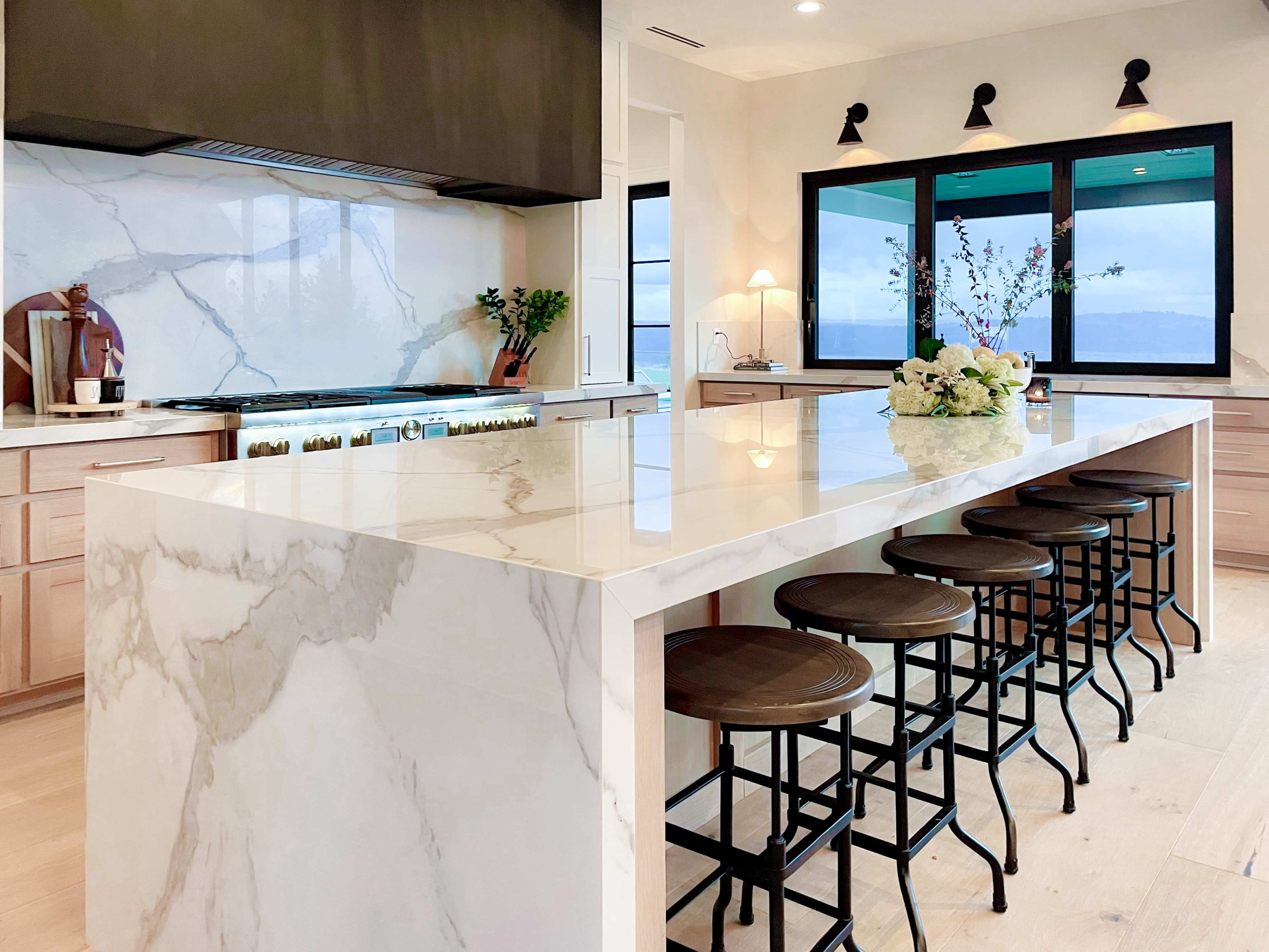 The image shows a modern kitchen featuring a marble island with bar stools, a stove, and large windows facing an outdoor view.