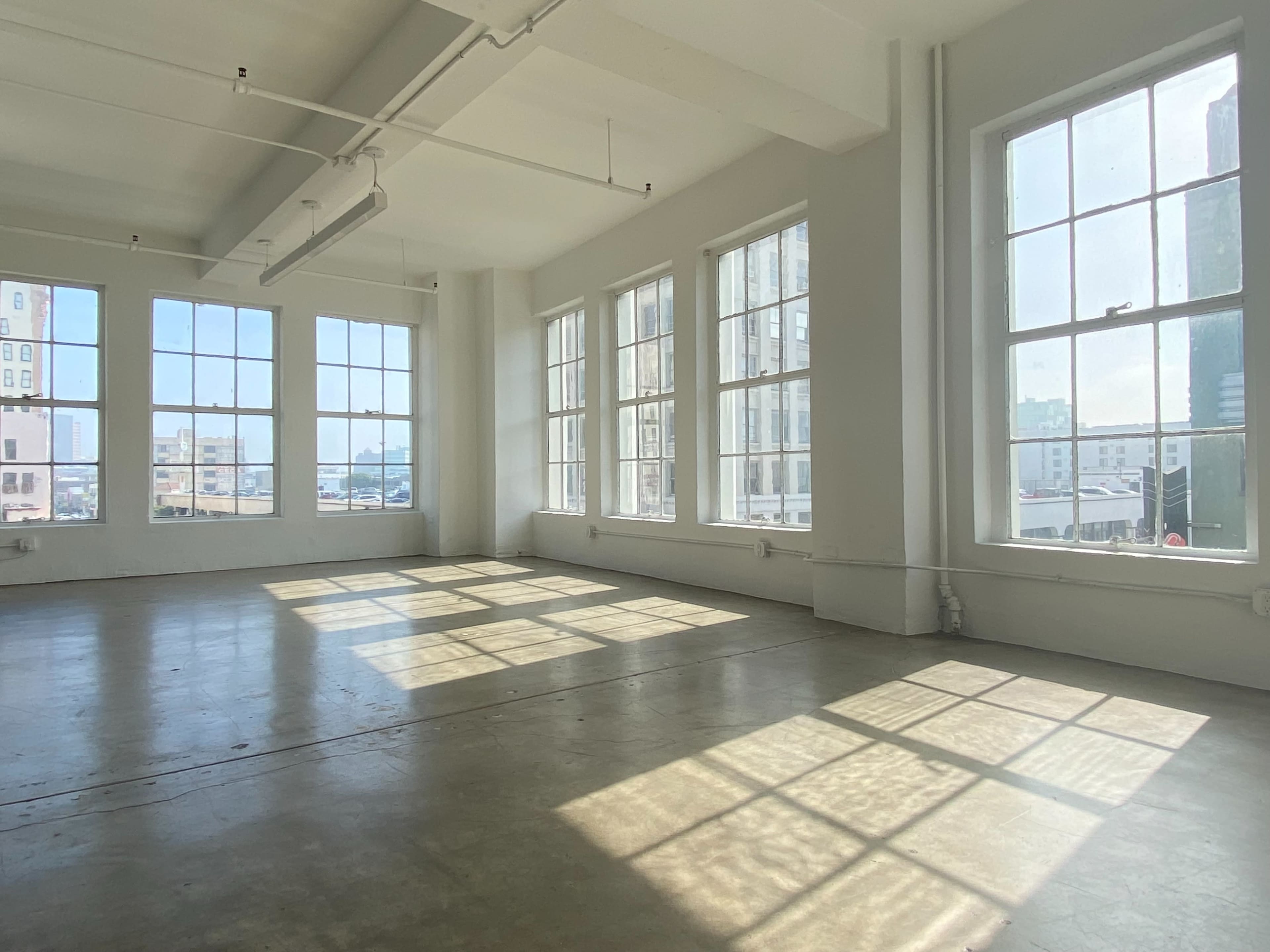 The image shows a spacious room with large windows allowing sunlight to cast shadows on the bare floor.