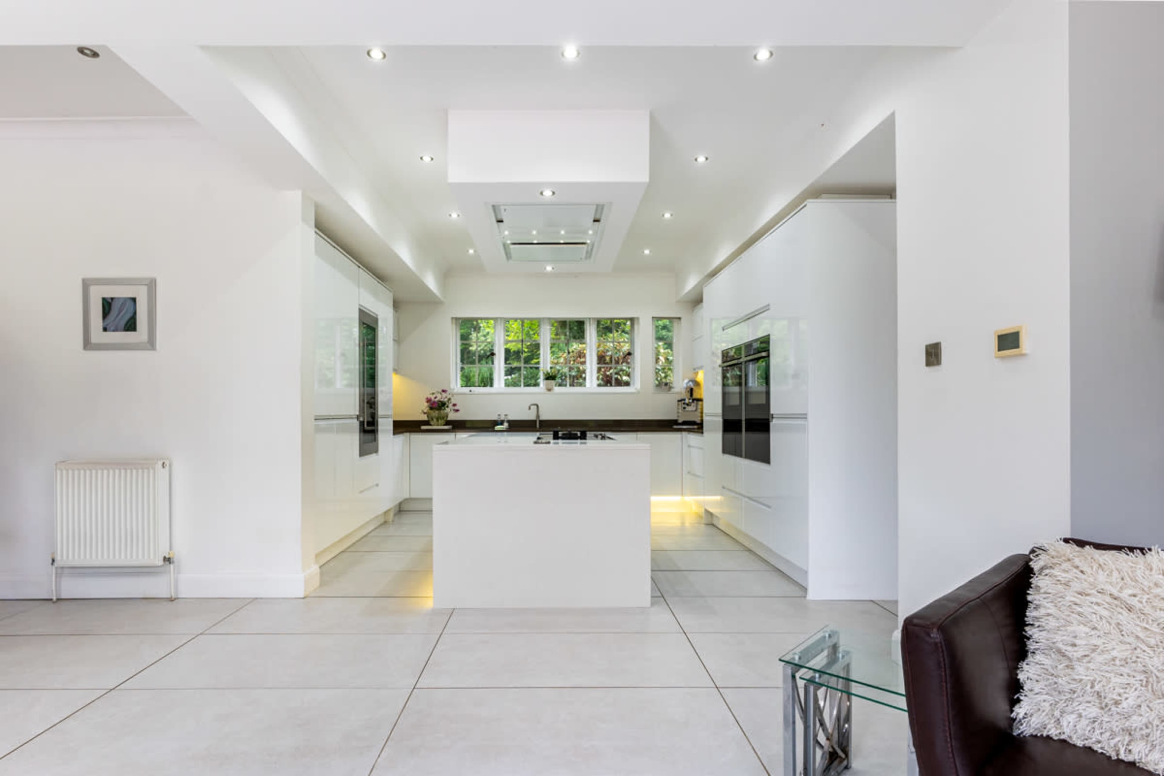 A modern kitchen features white cabinetry, a central island, and large tiles, all illuminated by recessed lighting.