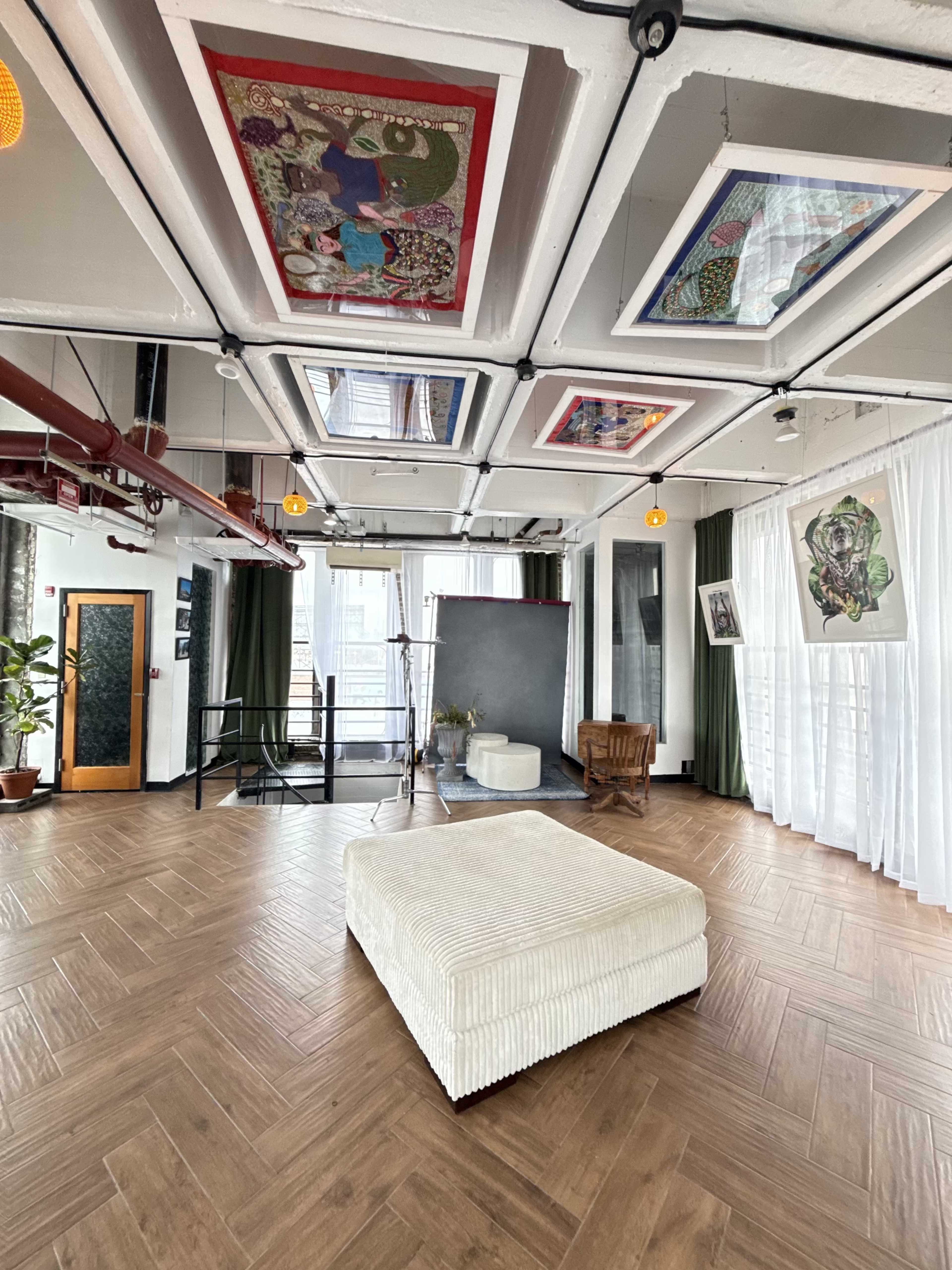 The image shows a spacious, modern room with a wooden floor, large windows, and artistic ceiling panels, featuring a central white ottoman and green curtains.