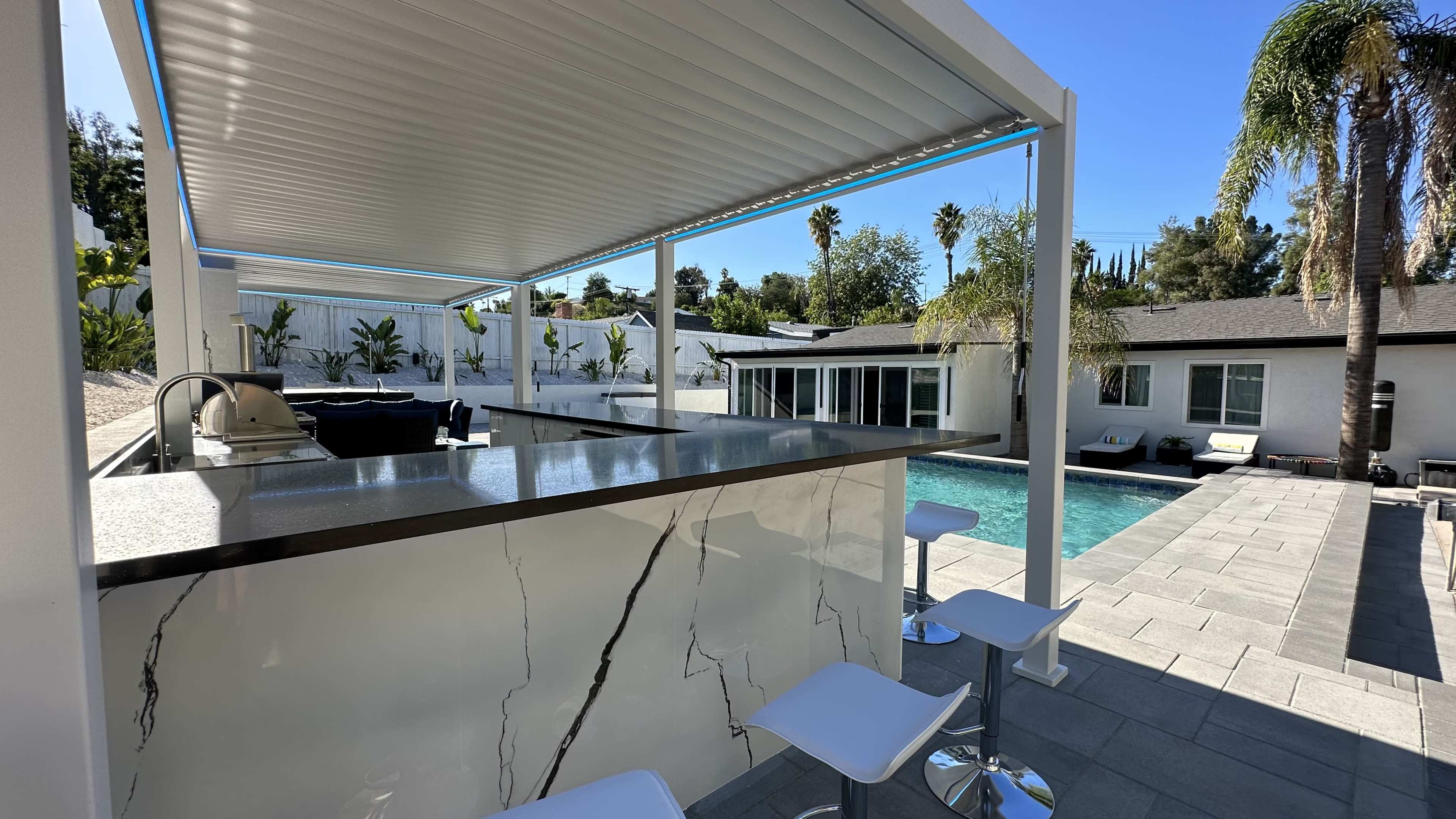 A modern outdoor bar area with a pool in the background, surrounded by palm trees and a white house.
