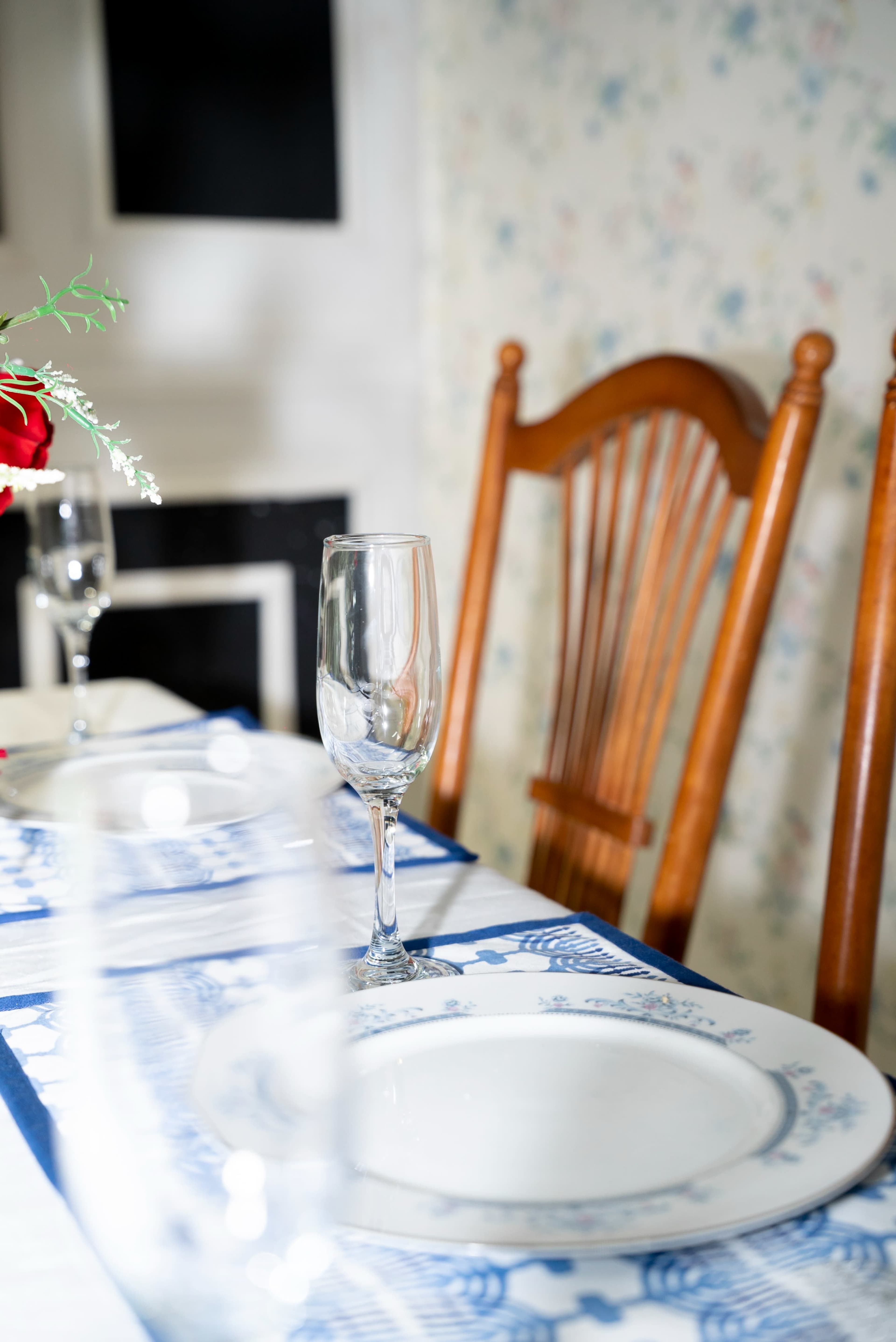 A dining table is set with white plates and a blue patterned tablecloth, accompanied by a wooden chair and a vase of red roses.