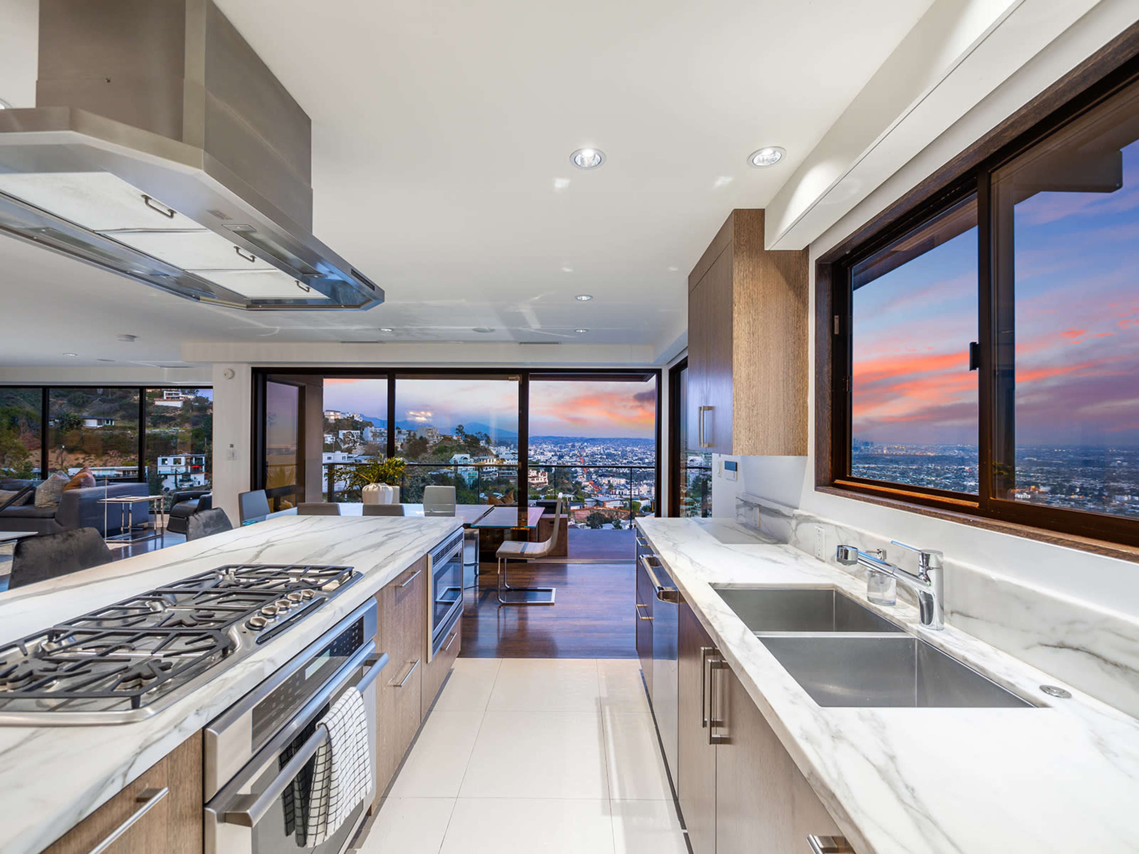The image shows a modern kitchen with a marble countertop, stainless steel appliances, and large windows offering a view of a sunset over a city.
