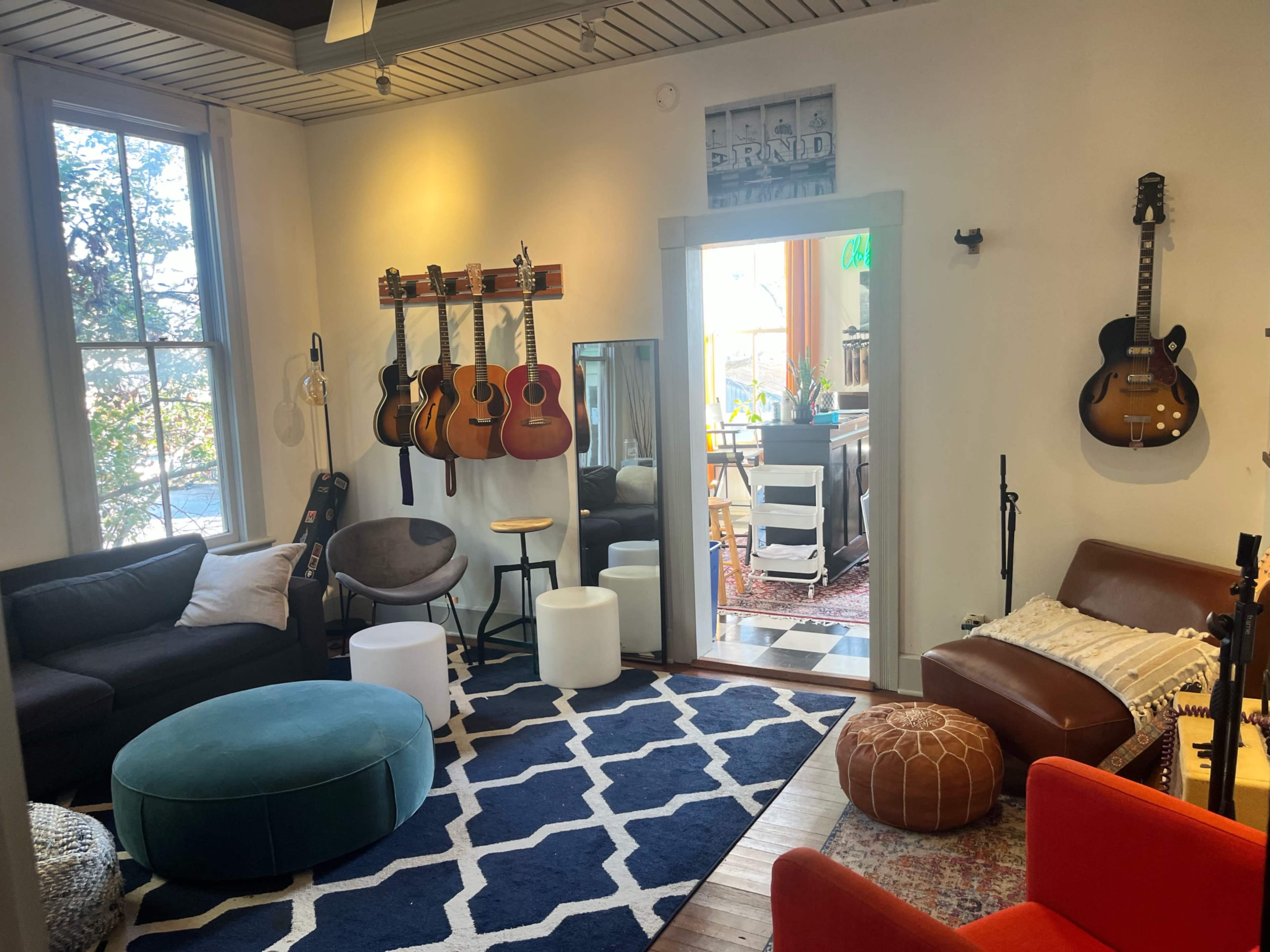 The image shows a cozy living room with a mix of seating options, guitars mounted on the wall, and an open doorway leading to a bright outdoor area.