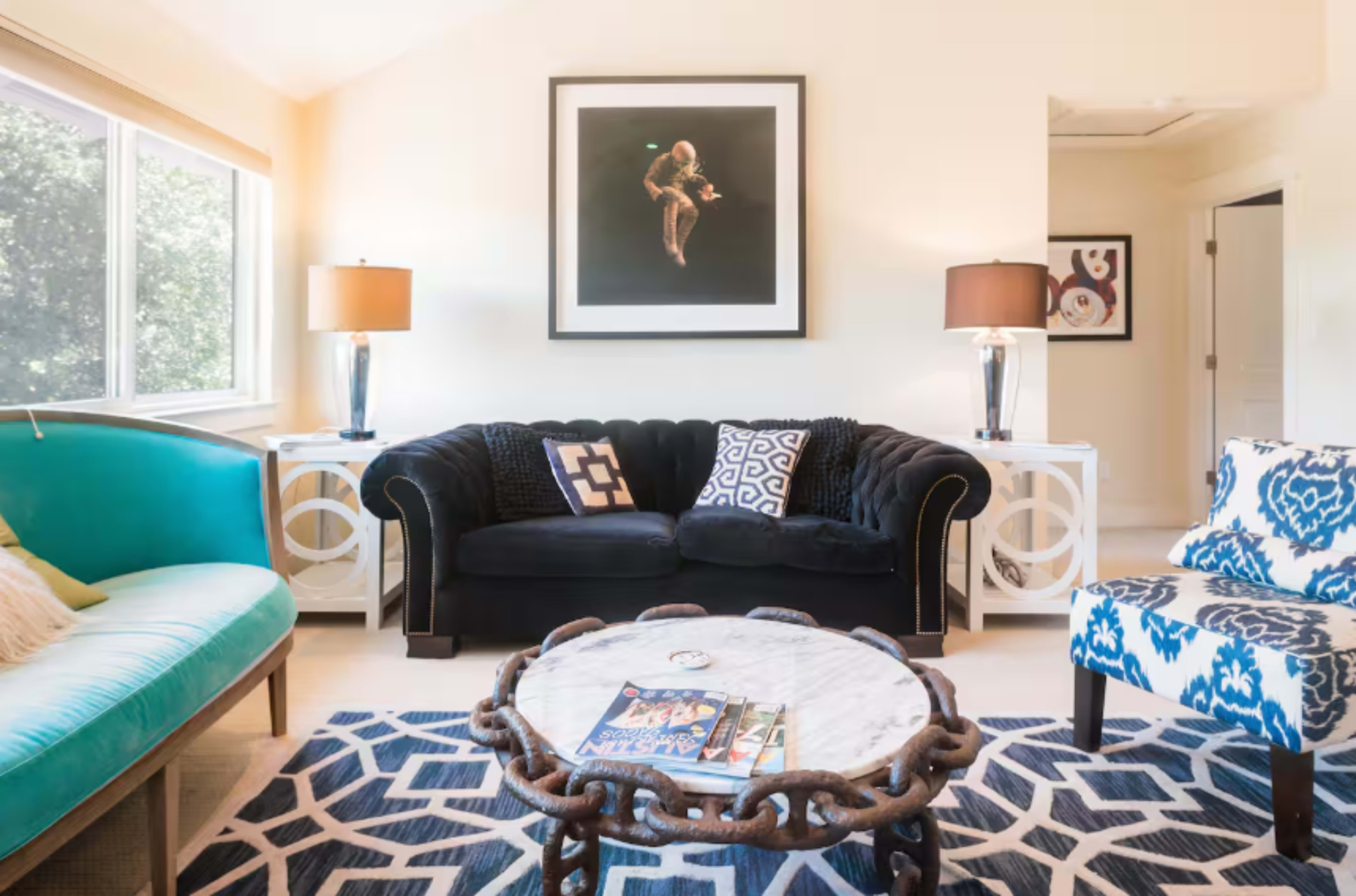 The image shows a brightly lit living room with a black sofa, a turquoise loveseat, a marble coffee table, and modern artwork on the walls.