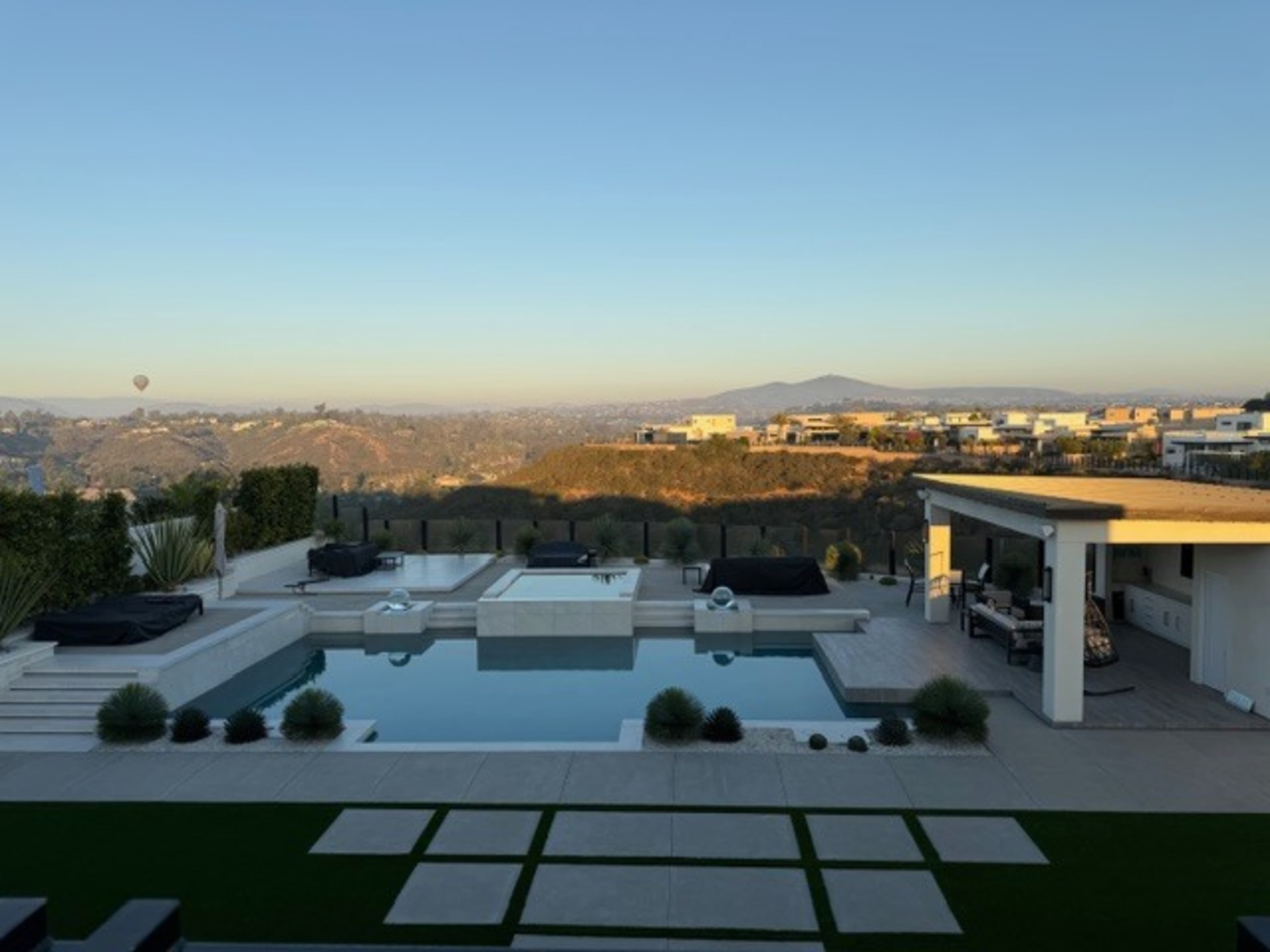 The image shows a modern outdoor pool area with a view of hills and distant homes, featuring lounge chairs and landscaped greenery.