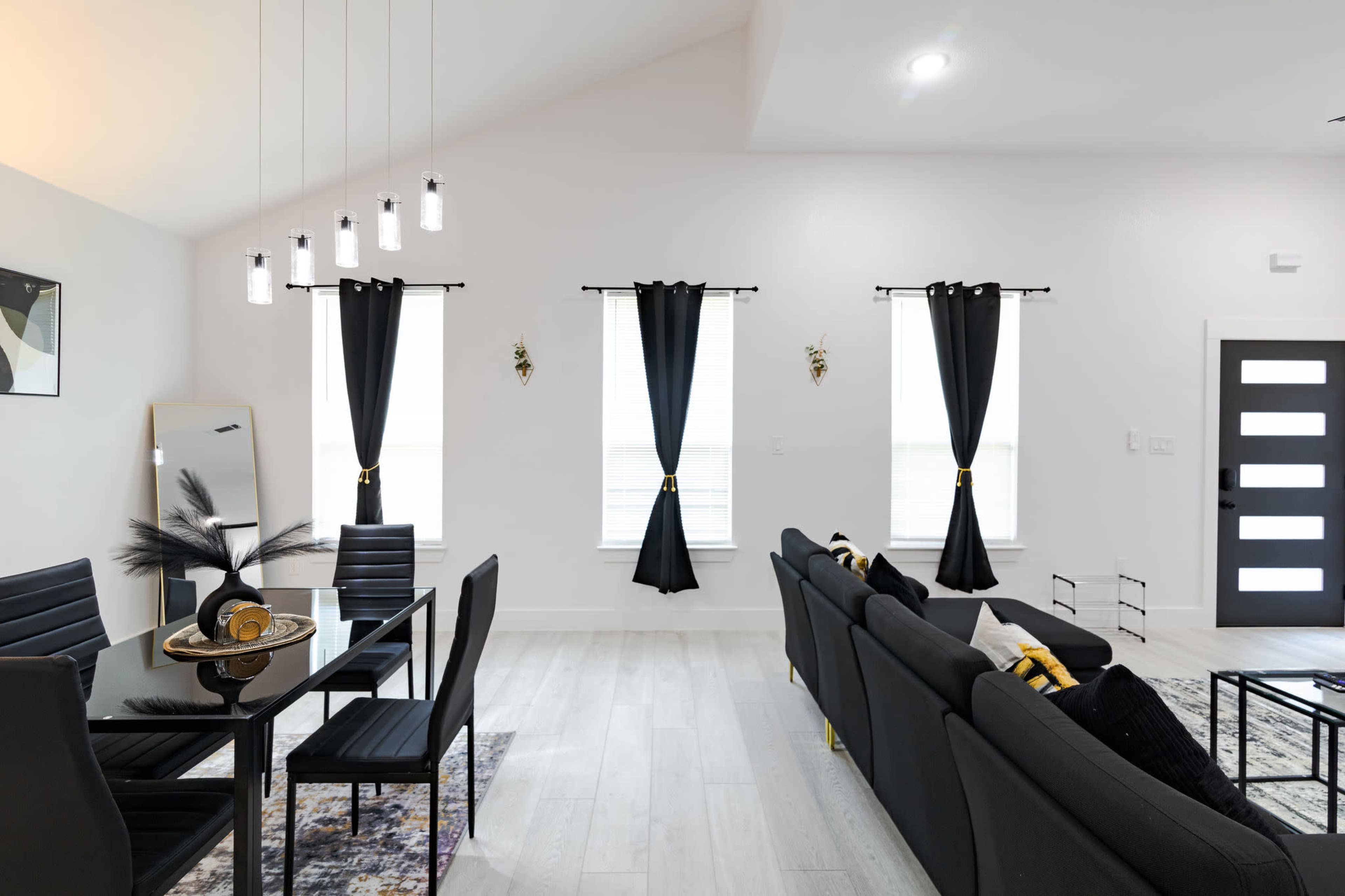 The image shows a modern living space featuring a black sectional sofa, a glass dining table with chairs, and large windows with black curtains.