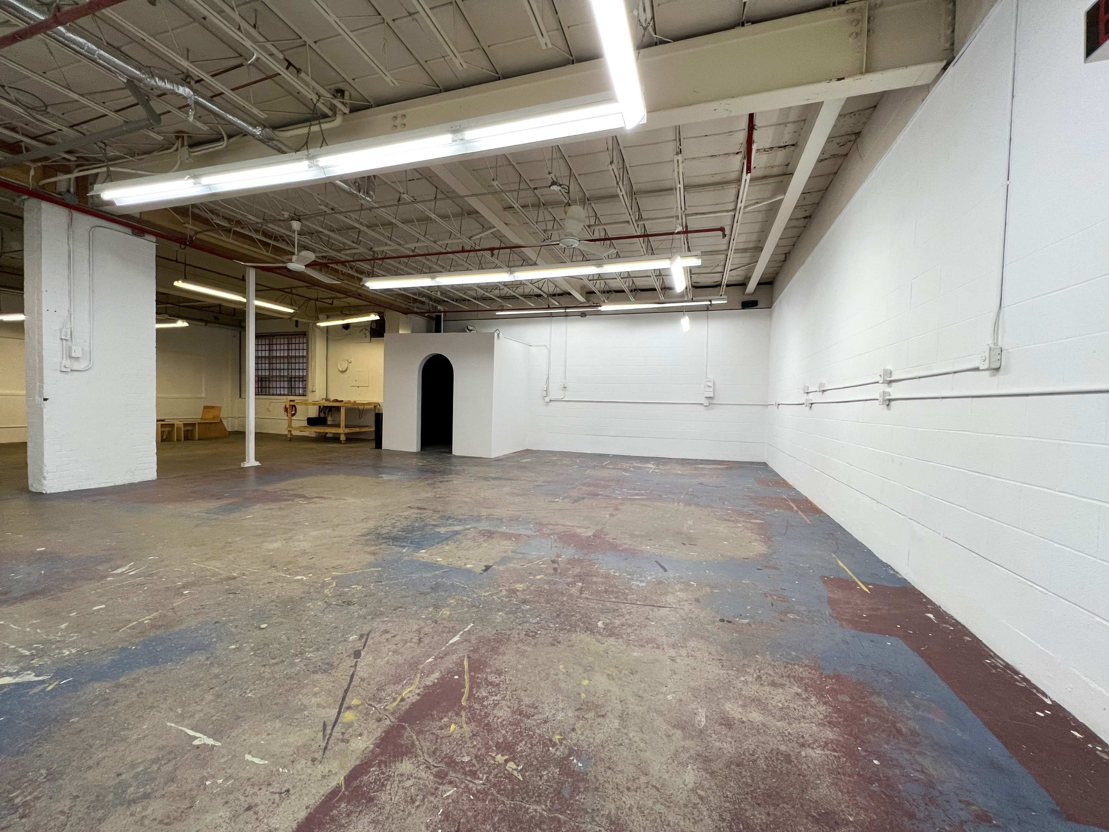 A spacious, empty warehouse with concrete floors and white-painted walls, featuring a dark archway on one side.