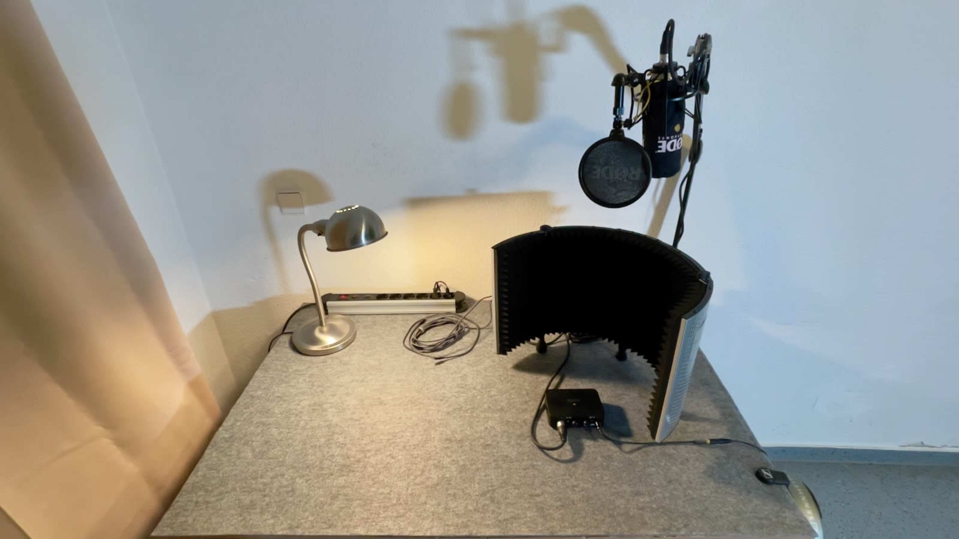 The image shows a workspace with a microphone setup, a soundproofing panel, a desk lamp, and audio equipment on a gray table.