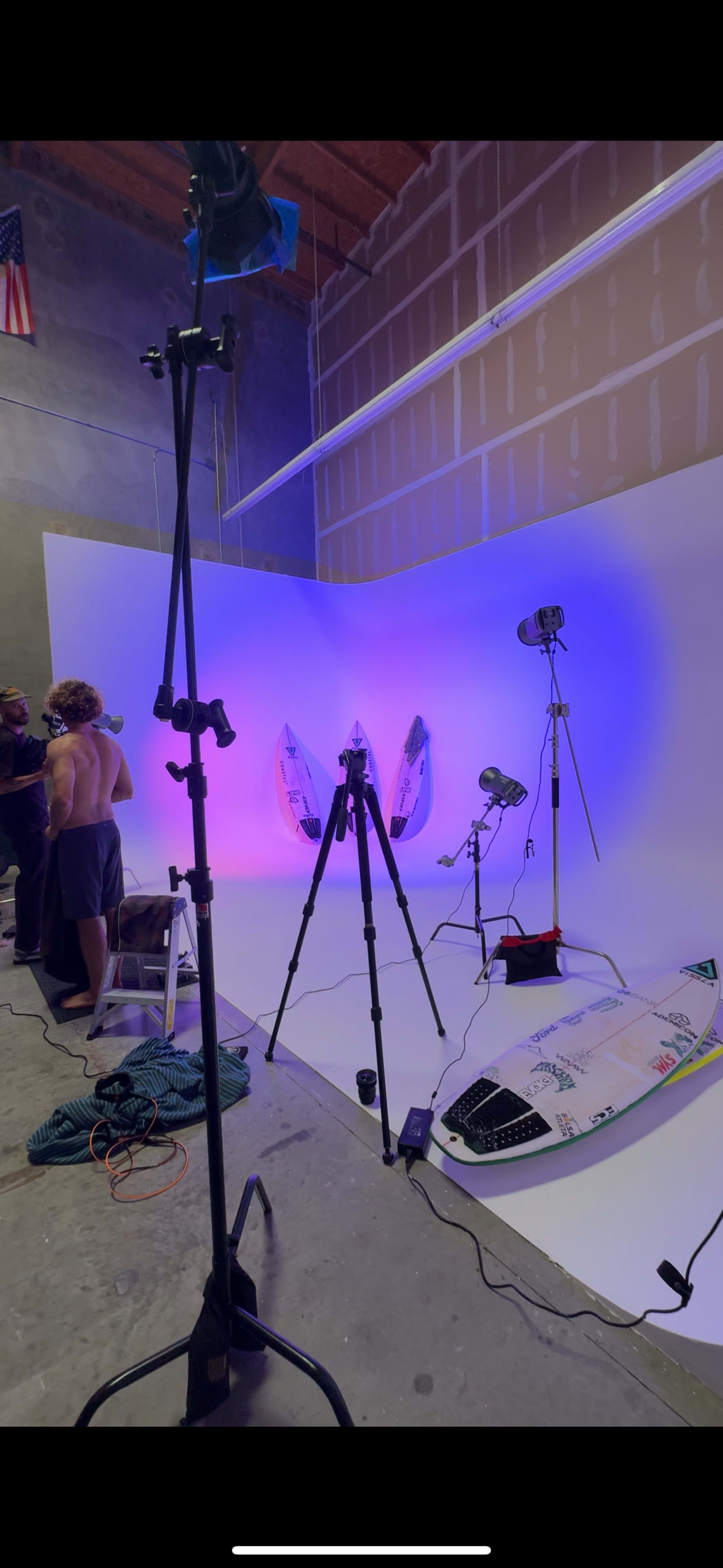 The image shows a photography studio setup with surfboards positioned against a colorful backdrop and various lighting equipment arranged around the scene.