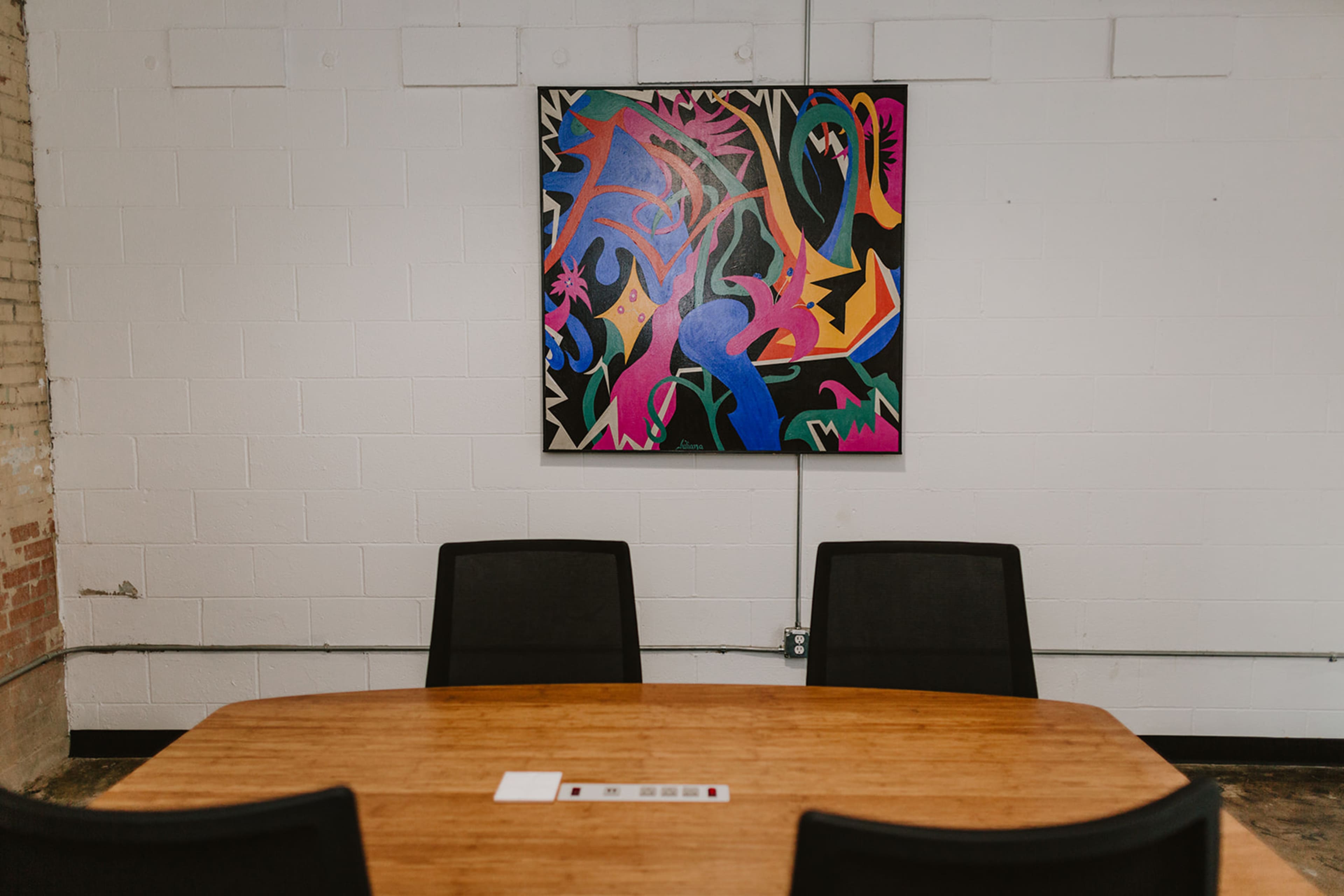 The image shows a conference room with a wooden table and three black chairs, featuring a colorful abstract painting on the white wall.