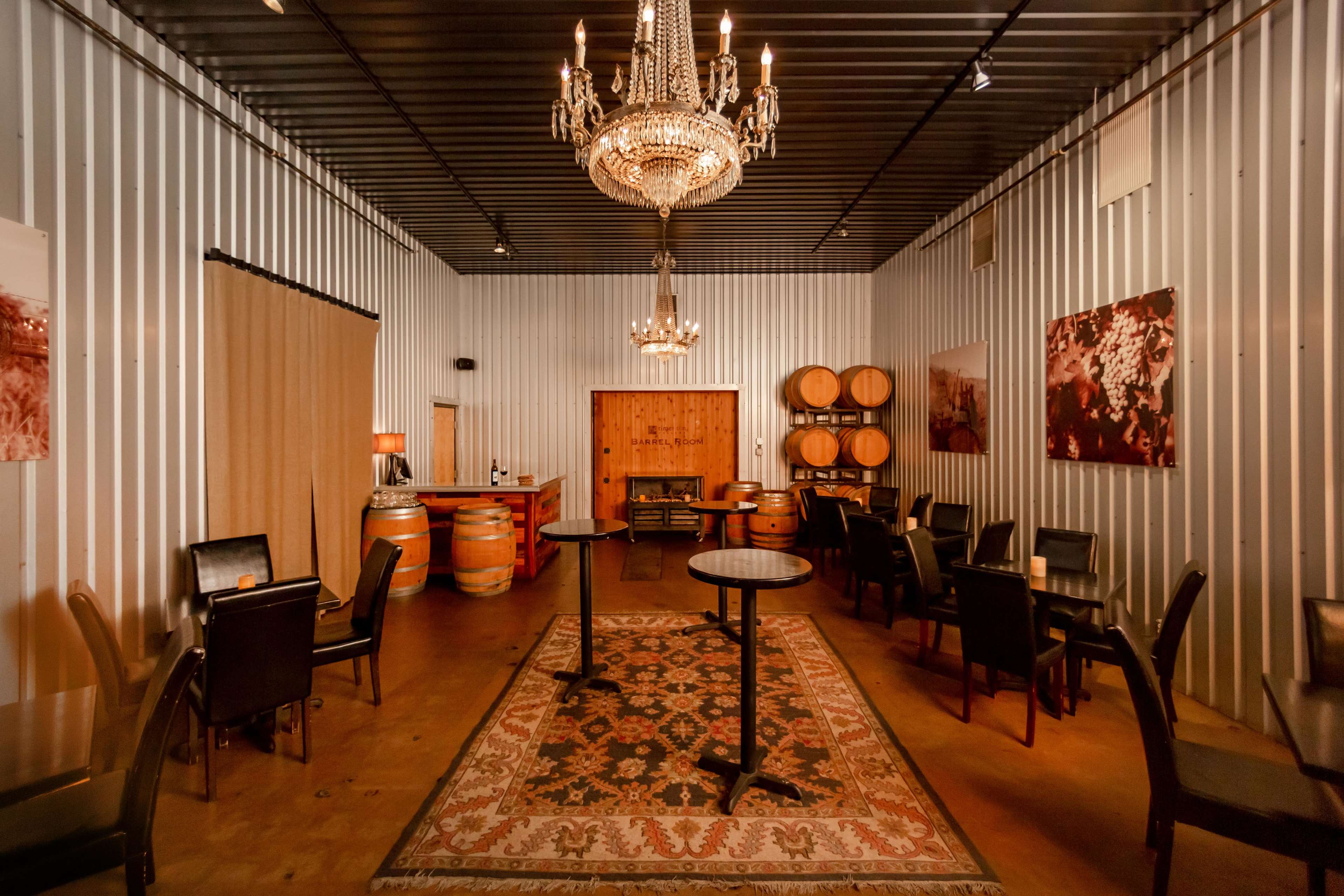 The image shows a dimly lit, metal-walled room furnished with tables, chairs, a chandelier, and wooden barrels.