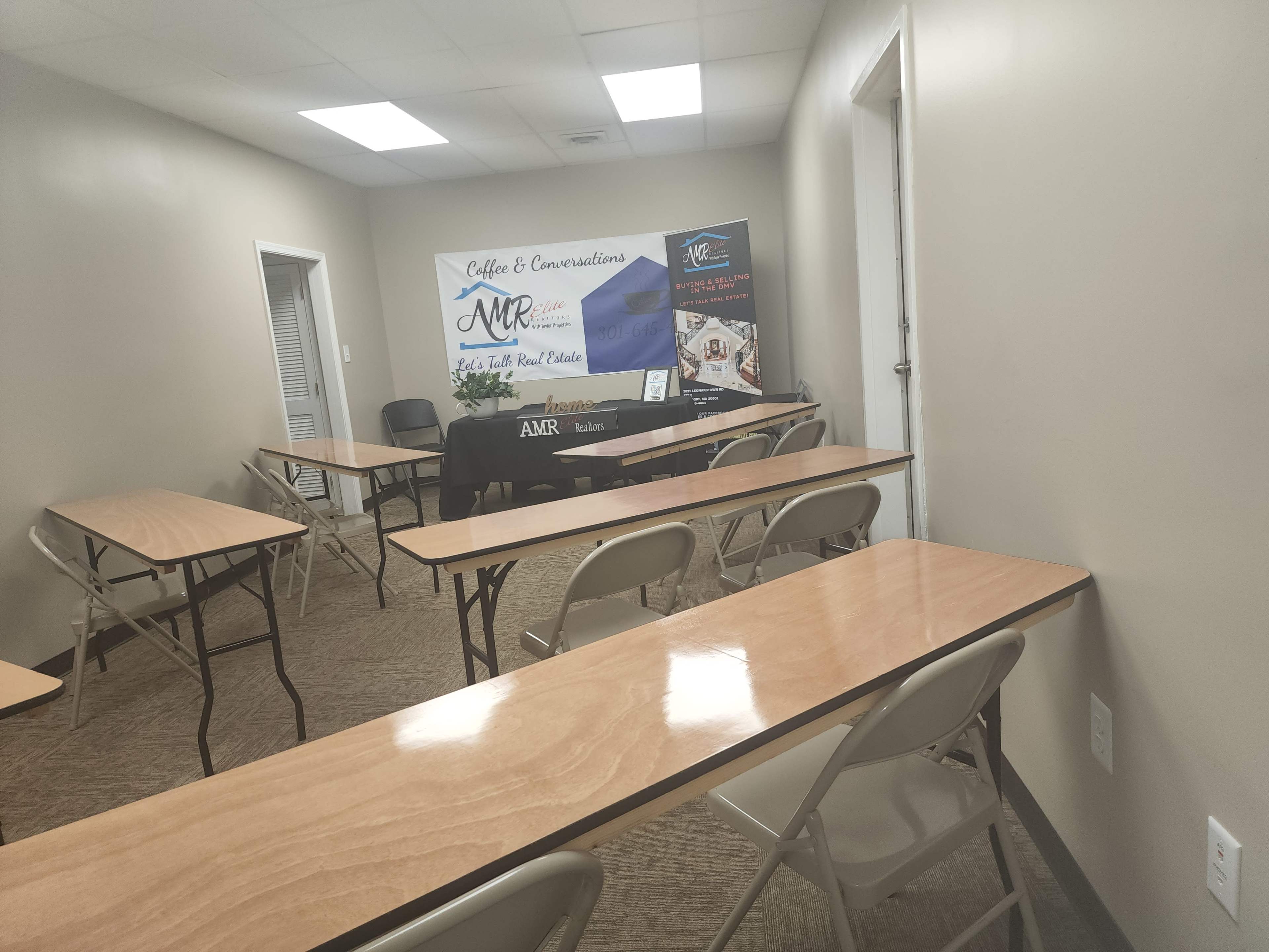 A sparsely furnished meeting room with several wooden tables and chairs arranged in a classroom style, featuring a promotional banner for "Coffee & Conversations" and "AMR" at the front.