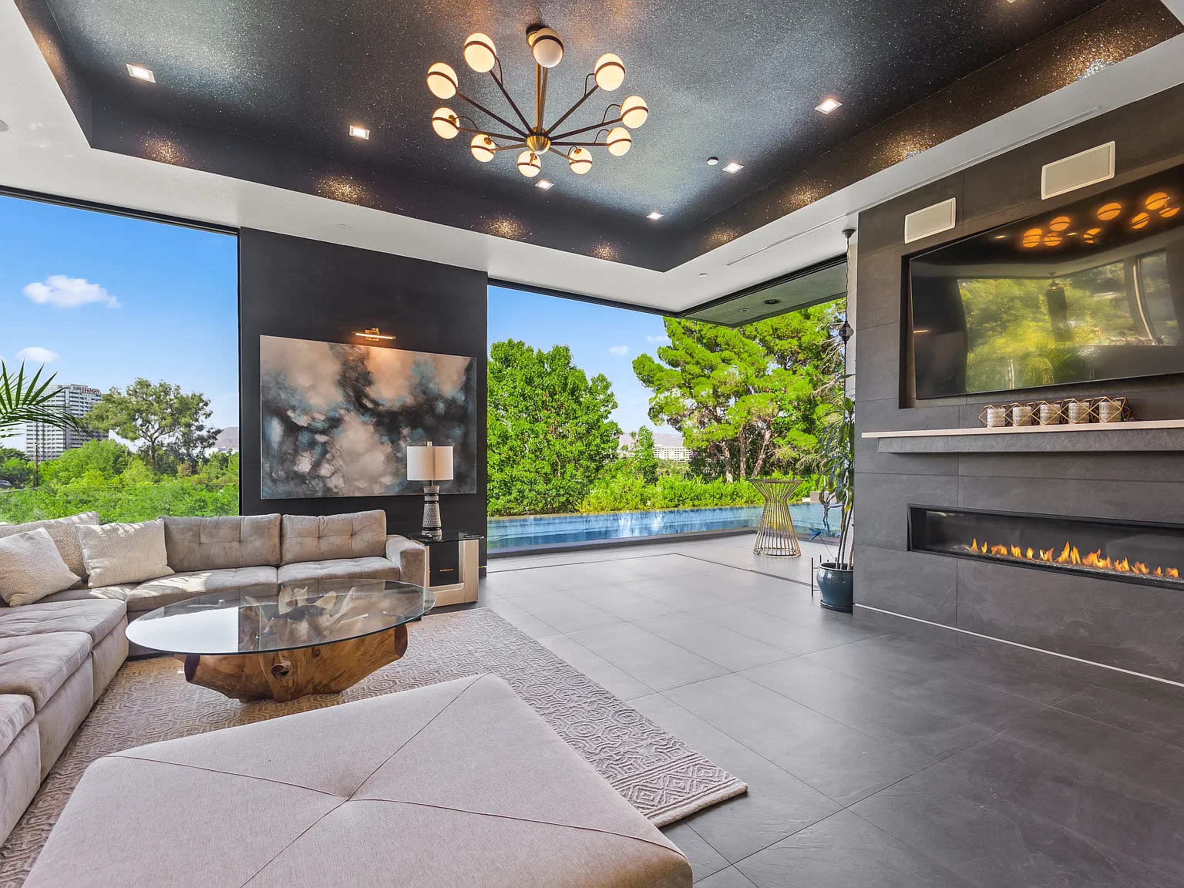 The image shows a modern living room with a large sectional sofa, a circular coffee table, and a wall-mounted television, featuring a large glass window that overlooks a pool and greenery outside.