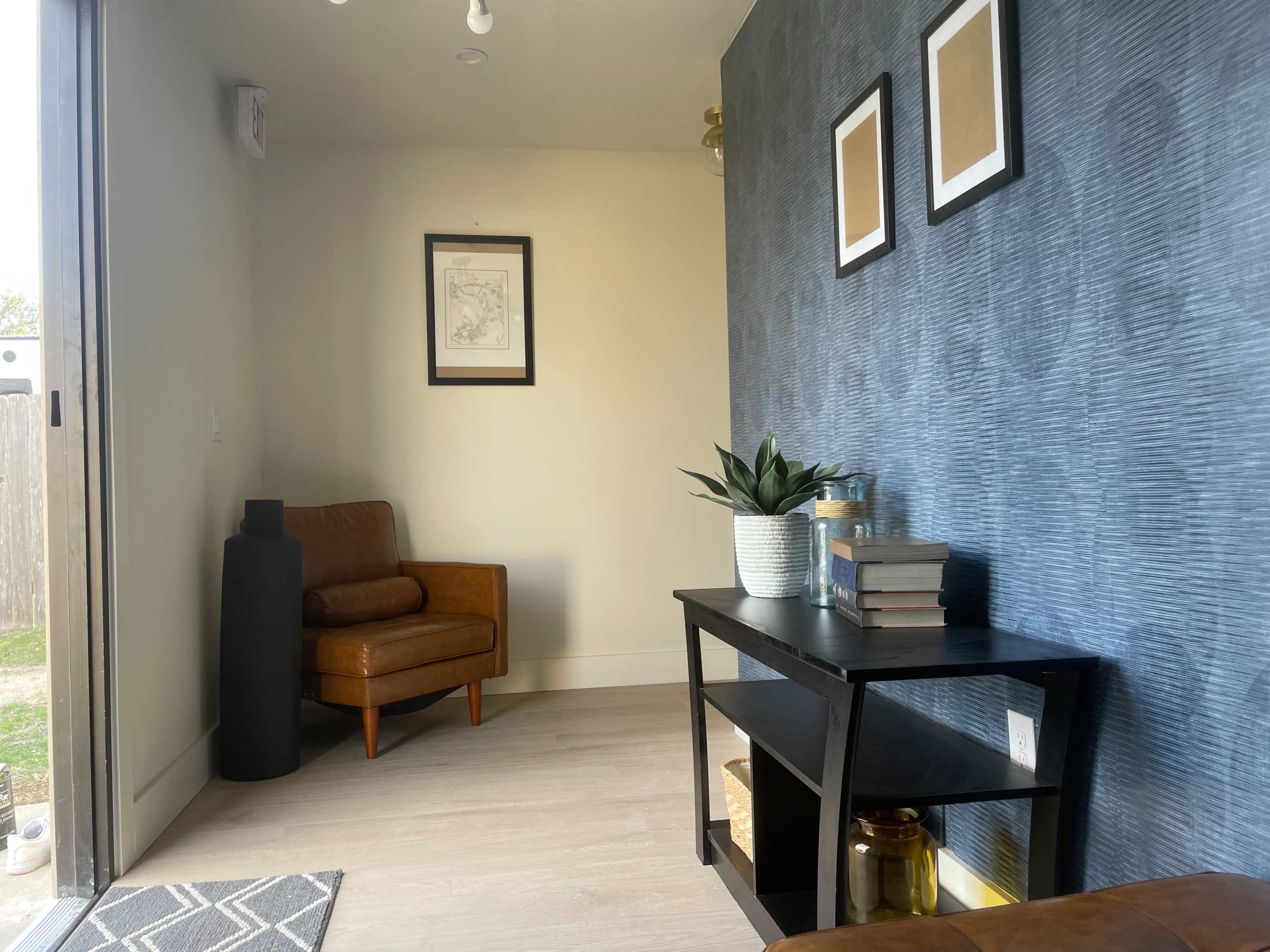 The image shows a small interior space featuring a brown leather chair, a black side table with books and a plant, and framed pictures on the wall.