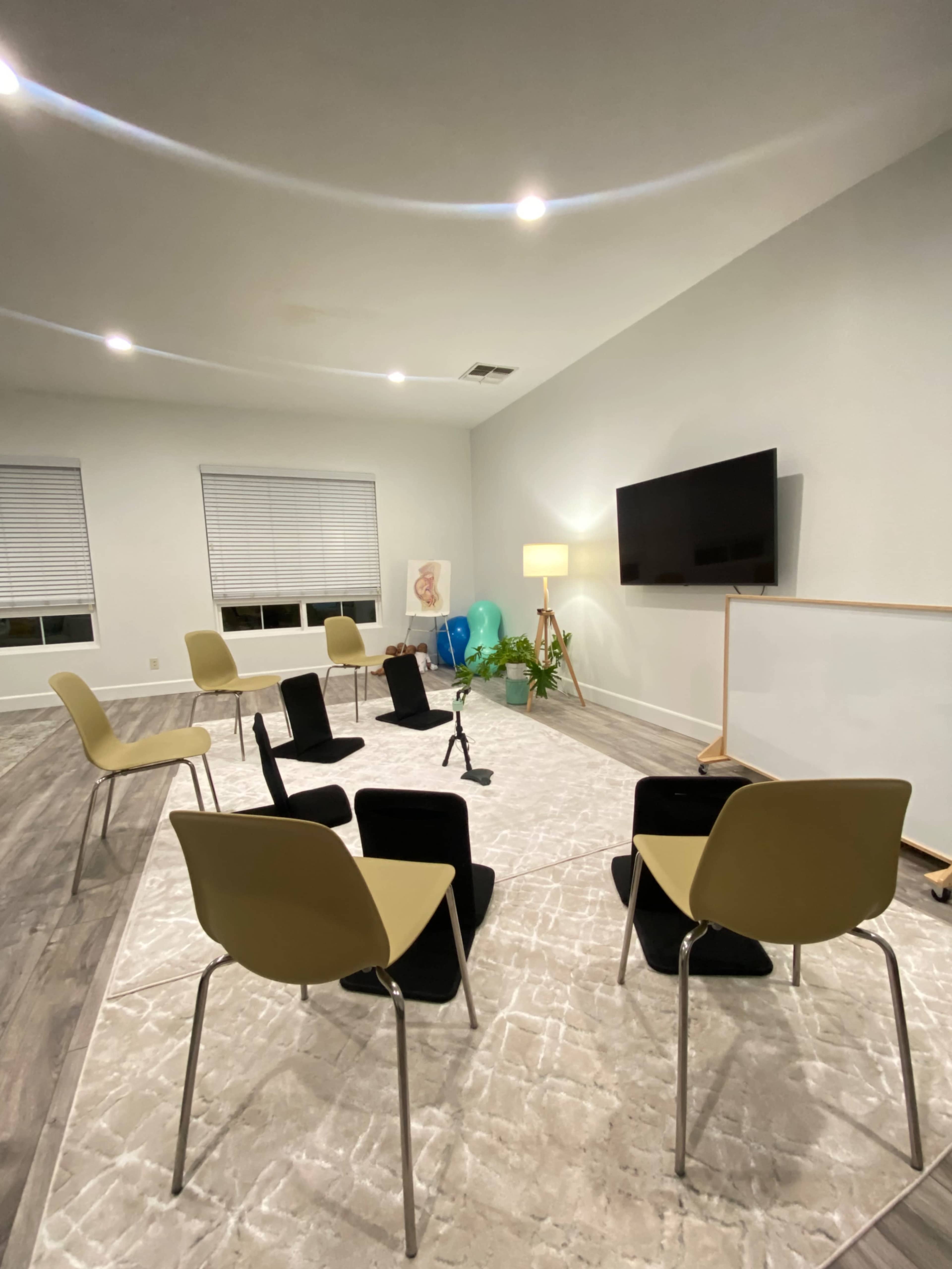 A room features six chairs arranged in a circle on a light-colored rug, with a television and whiteboard on opposing walls.
