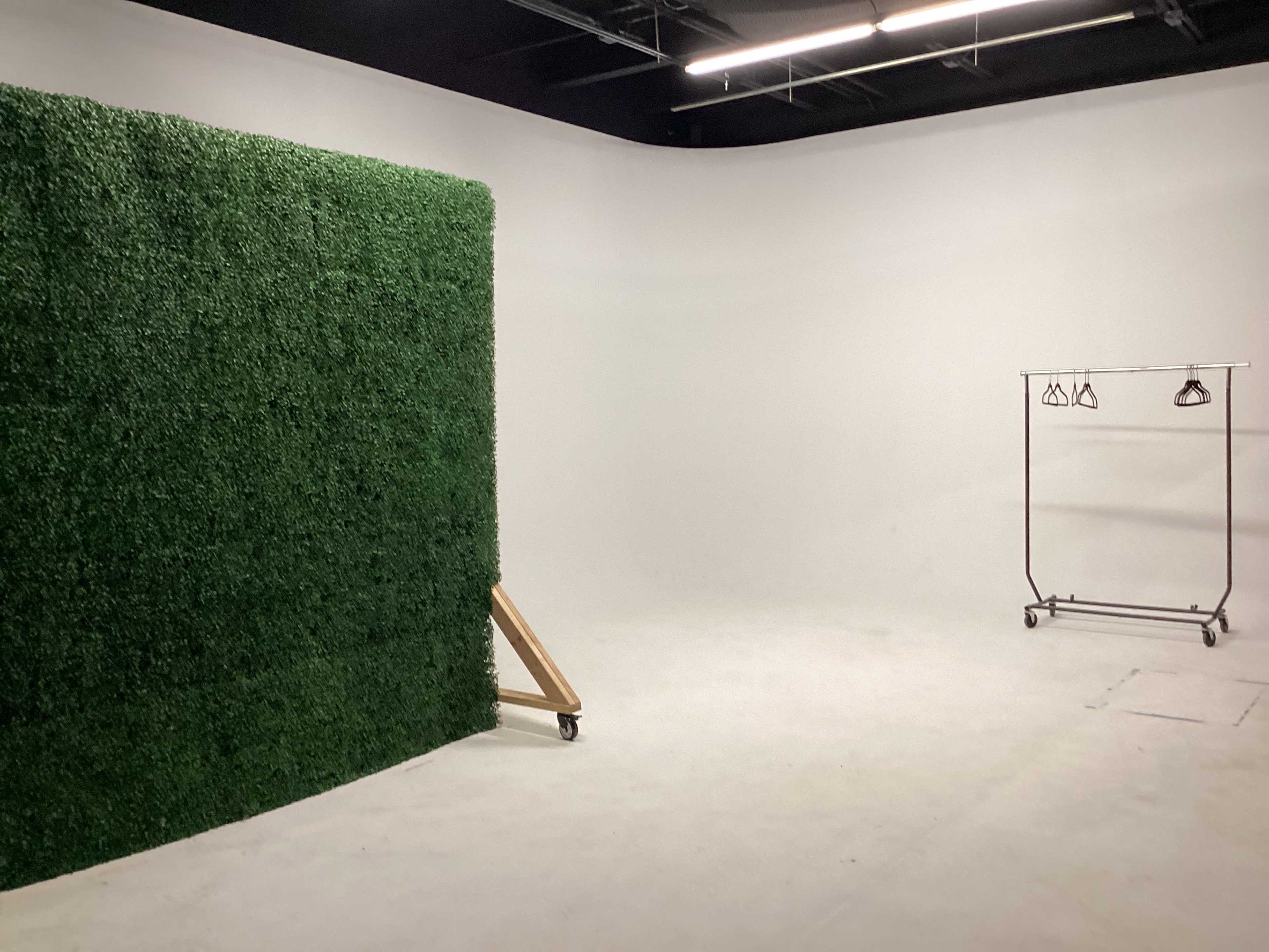 A minimalistic studio space with a green artificial grass backdrop on the left and a clothing rack with hangers on the right.