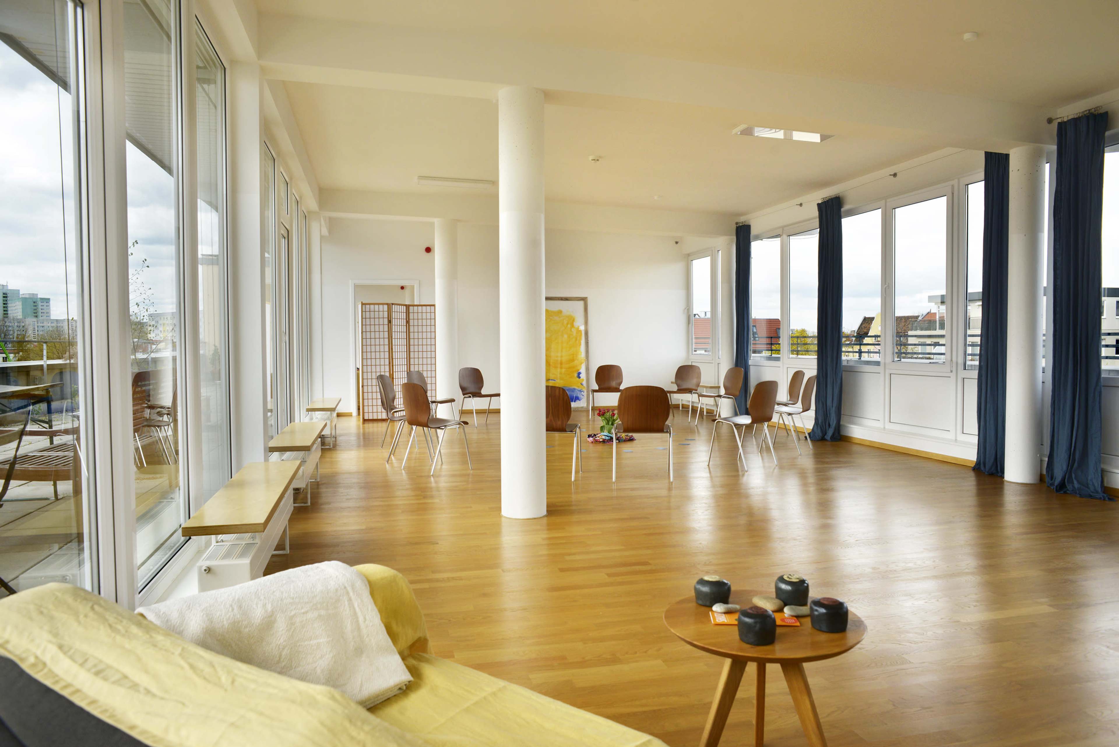 A spacious, well-lit room with wooden flooring, featuring a seating area with chairs arranged in a circle and large windows providing natural light.