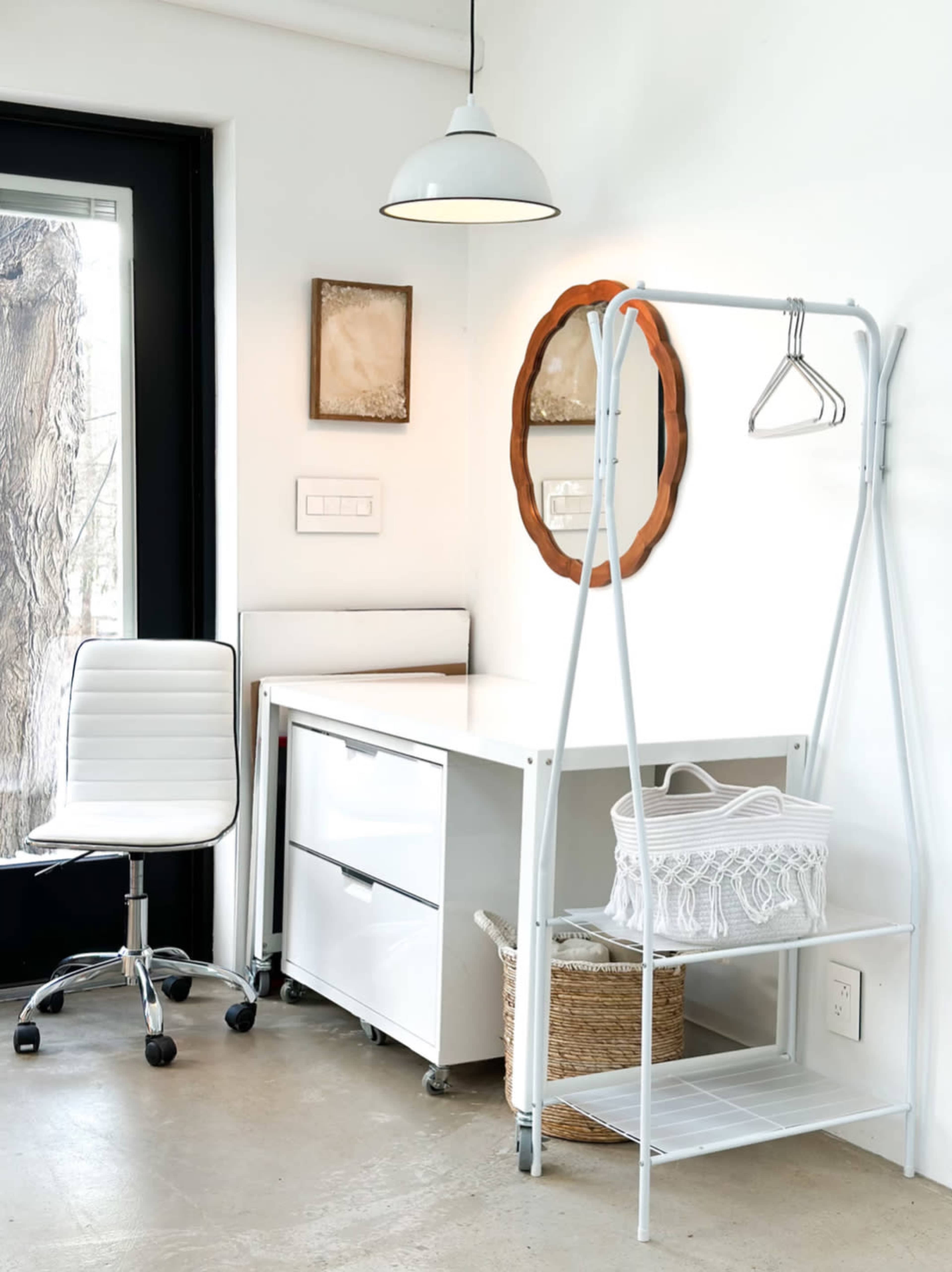 A minimalist workspace features a white desk, a swivel chair, a hanging clothing rack, and a decorative mirror on the wall.