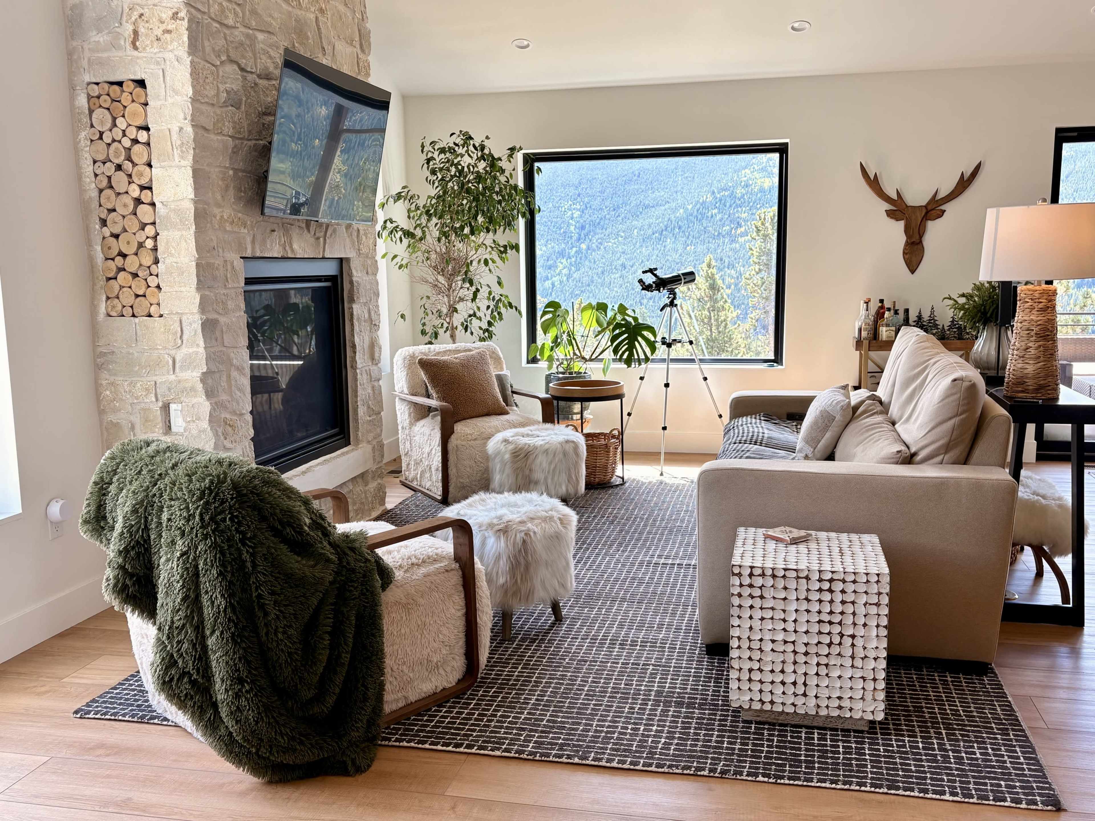 The image shows a modern living room with a stone fireplace, comfortable seating, large windows overlooking mountains, and decorative elements including a plant and a telescope.