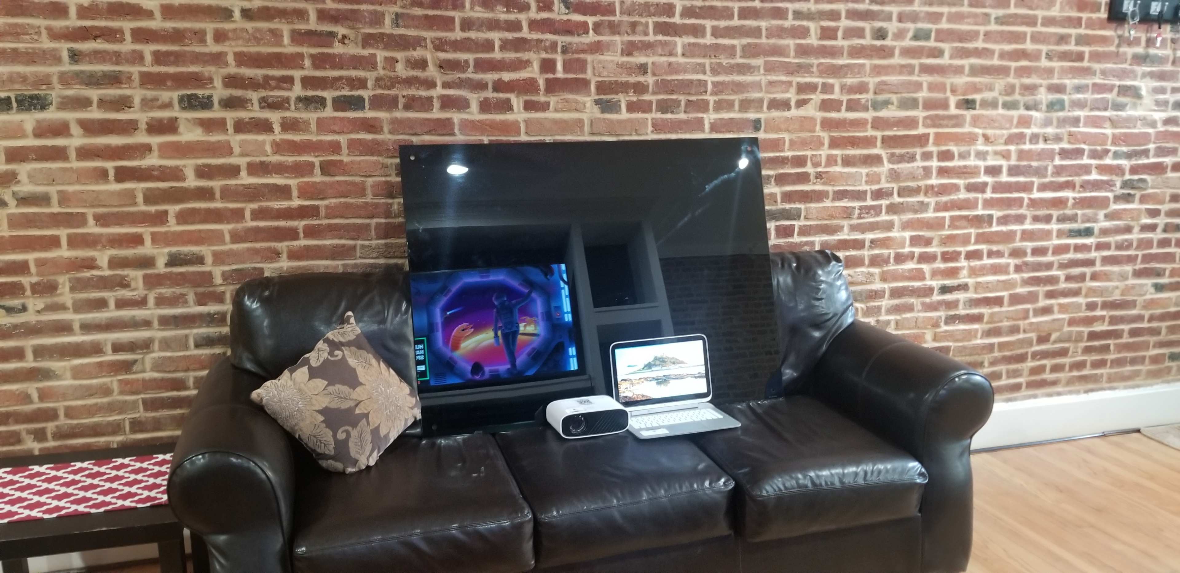 A black television is propped against a brick wall, with a laptop and a projector placed on a leather couch.