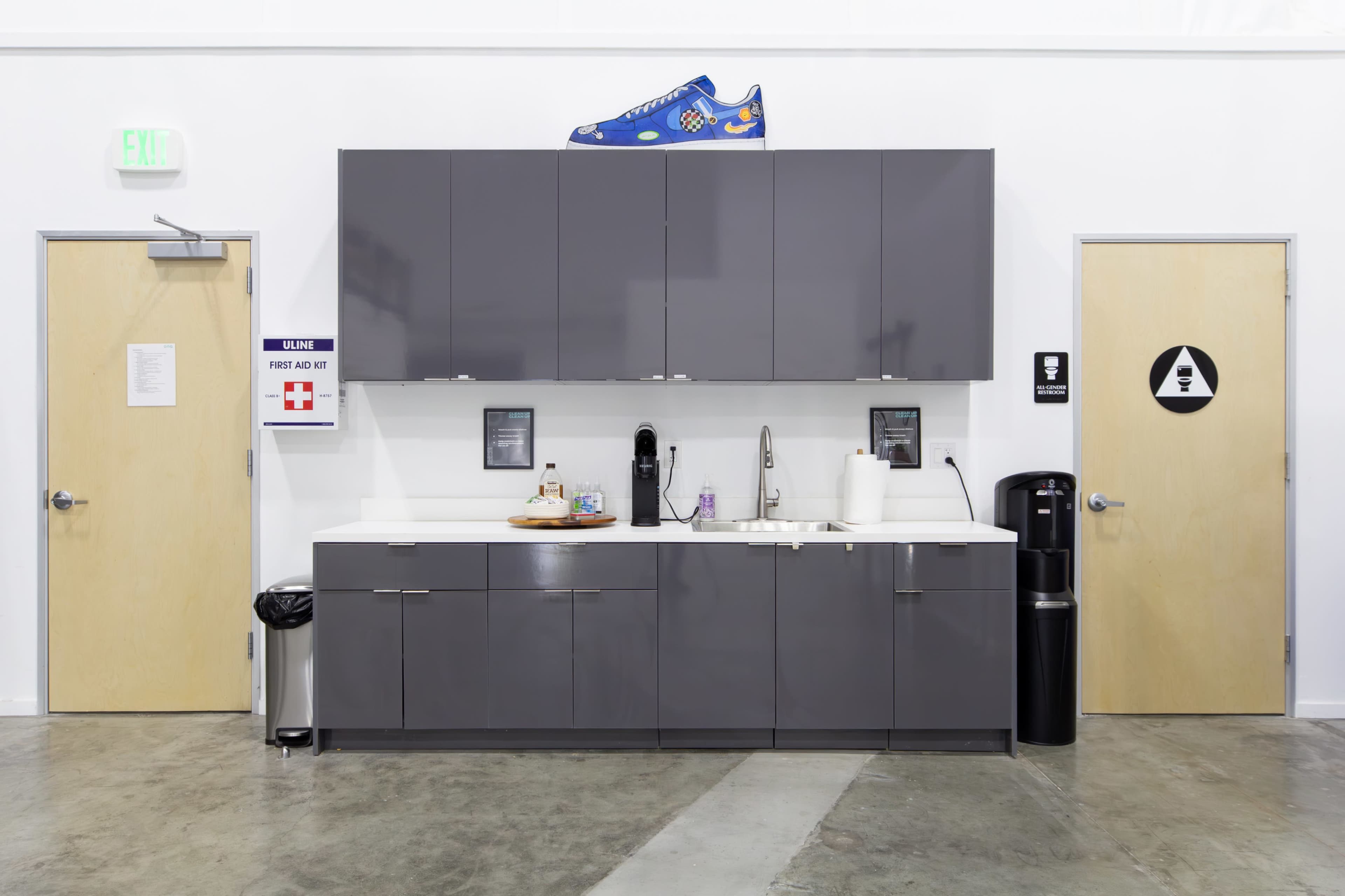 The image shows a modern break room with gray cabinets, a sink, a coffee machine, and a large blue sneaker displayed on top of the cabinets.