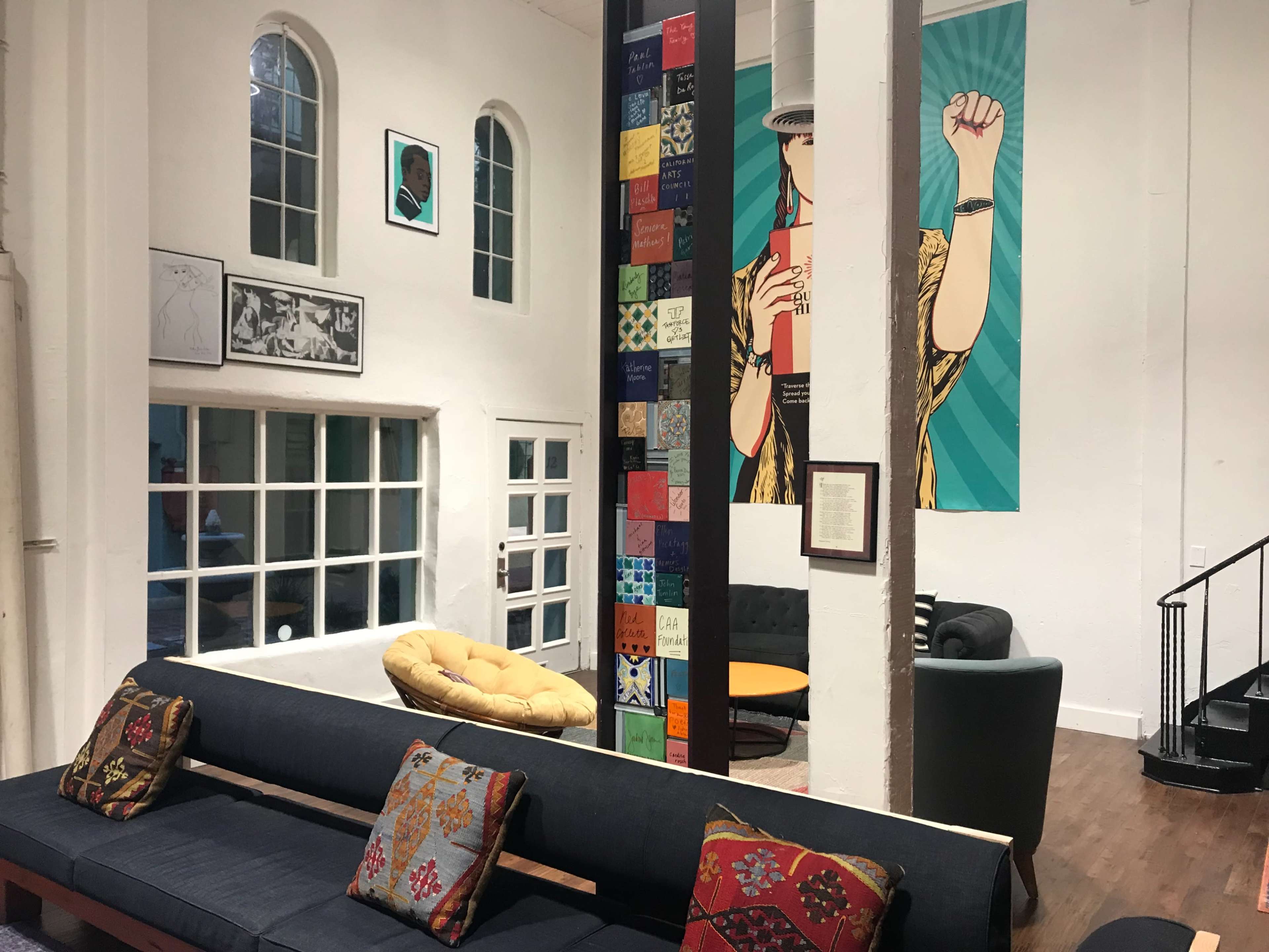 The image shows a spacious interior with a large seating area featuring colorful cushions, a prominent mural on the wall, and a staircase leading to an upper level.