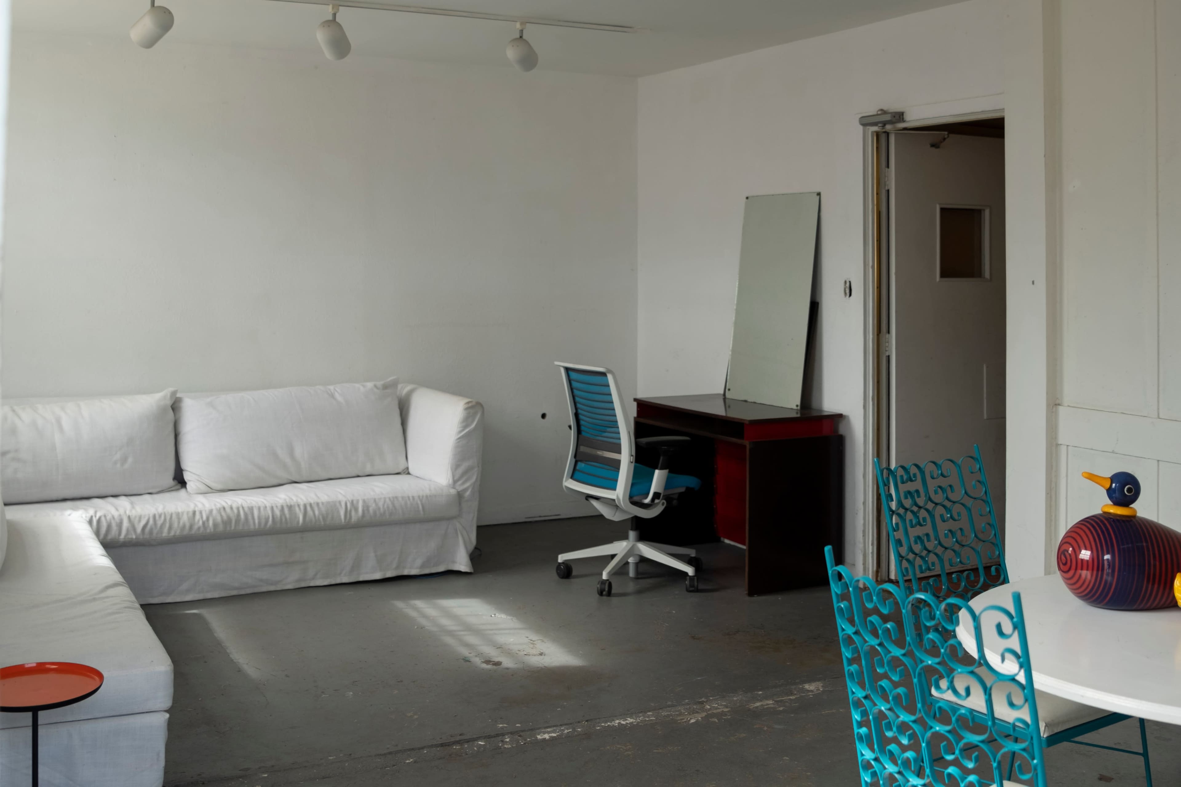 The image shows a spacious, minimalist room with a white sofa, a blue office chair at a desk, a mirror, and a colorful decorative item on the table.