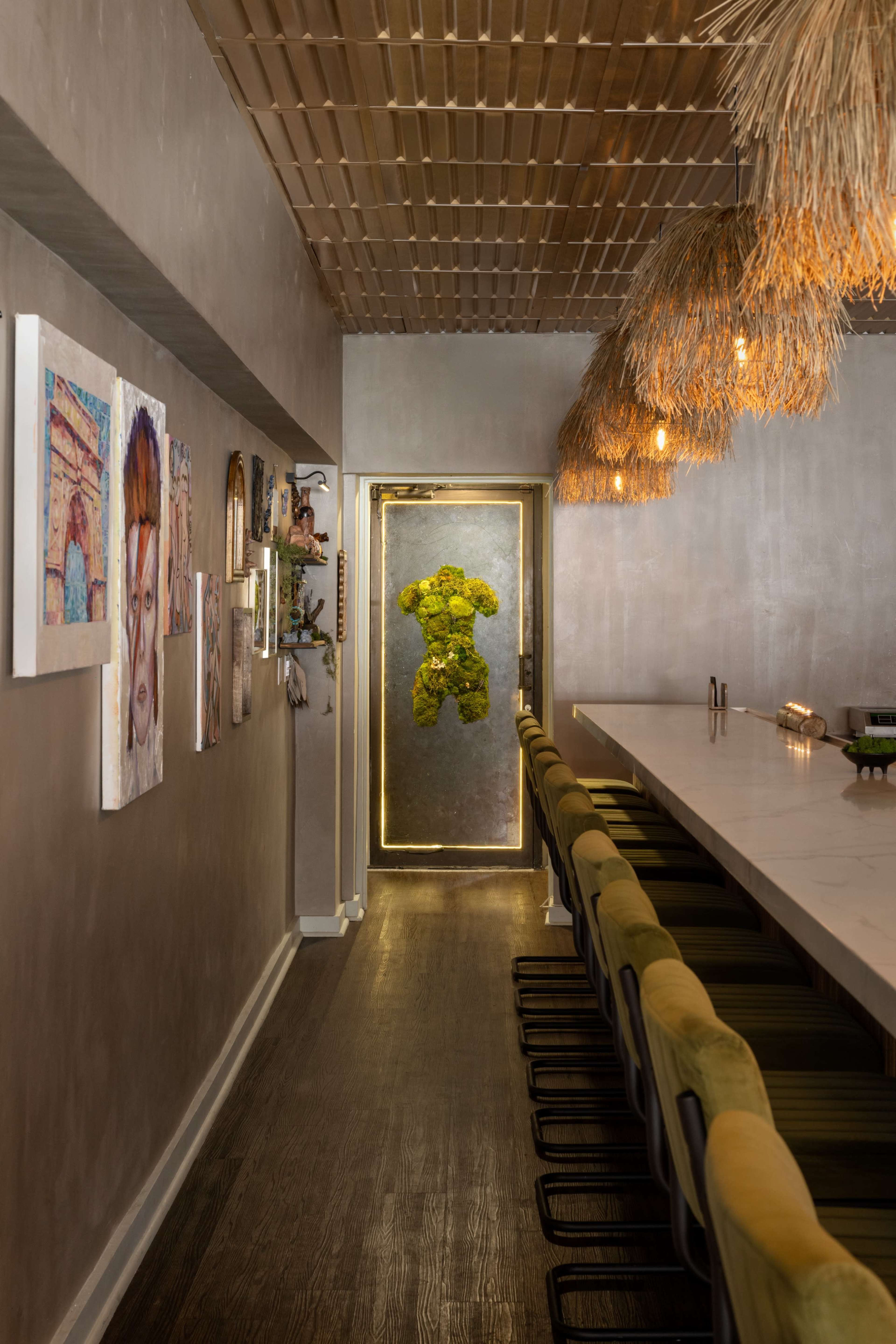 The image shows a narrow restaurant corridor featuring artwork on the walls, a long bar with green stools, and a distinctive door adorned with moss.