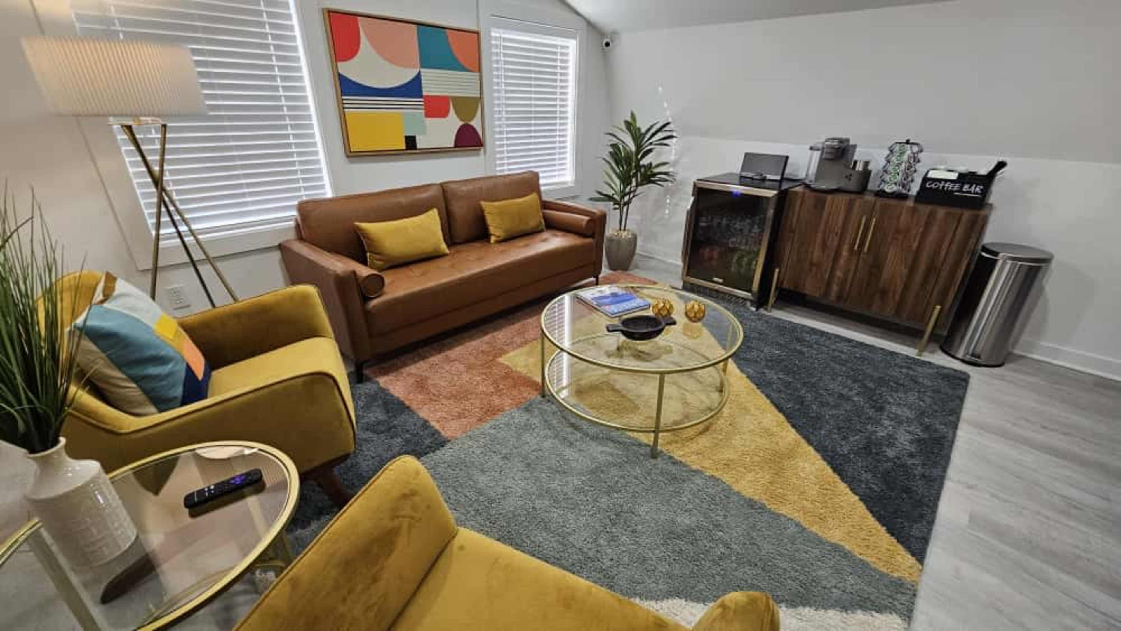 The image shows a modern living room with a brown leather sofa, yellow accent chairs, a circular coffee table, and a wooden sideboard against a wall adorned with colorful artwork.