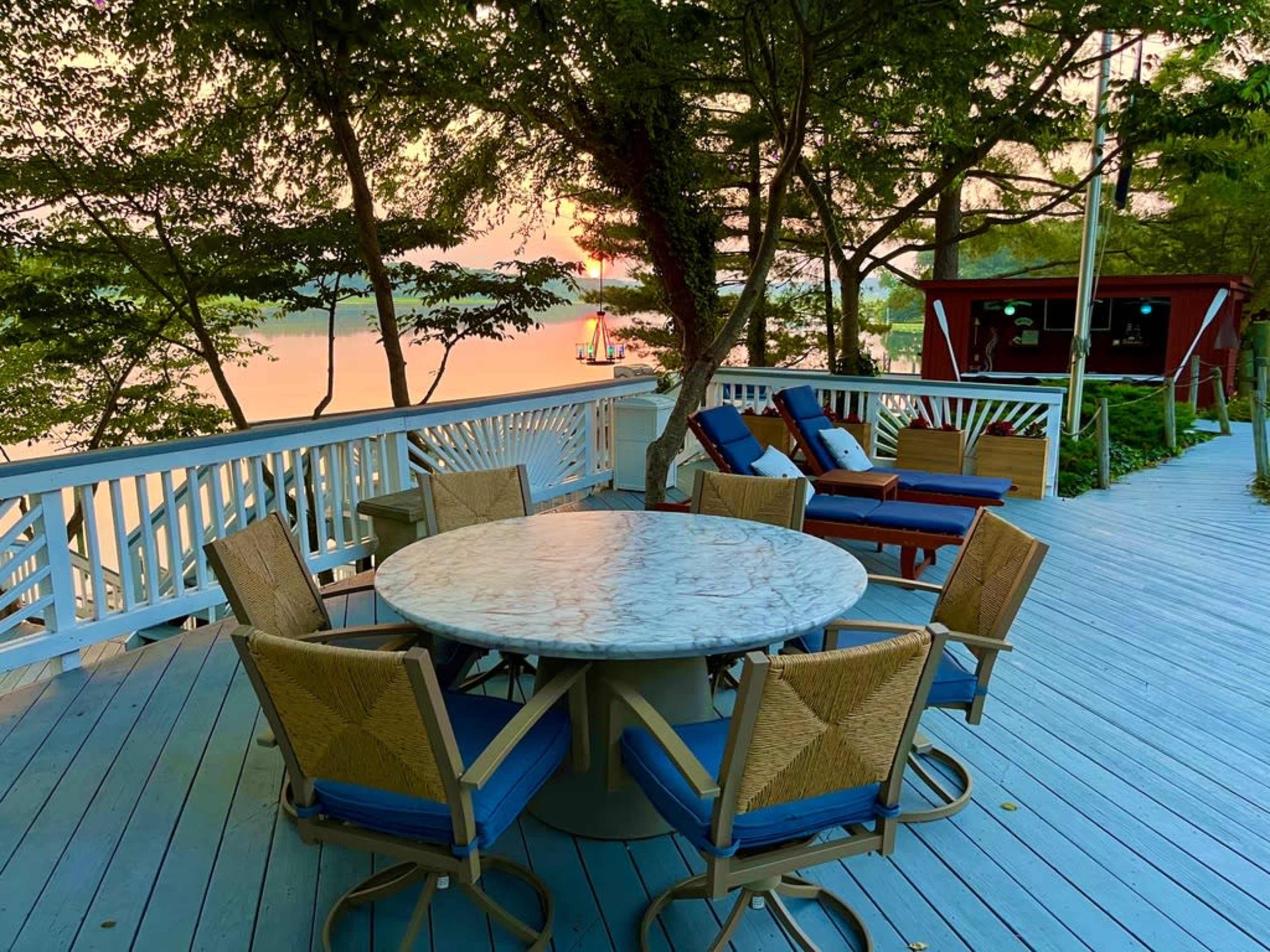 A circular marble table surrounded by eight chairs is set on a wooden deck overlooking a tranquil body of water at sunset.