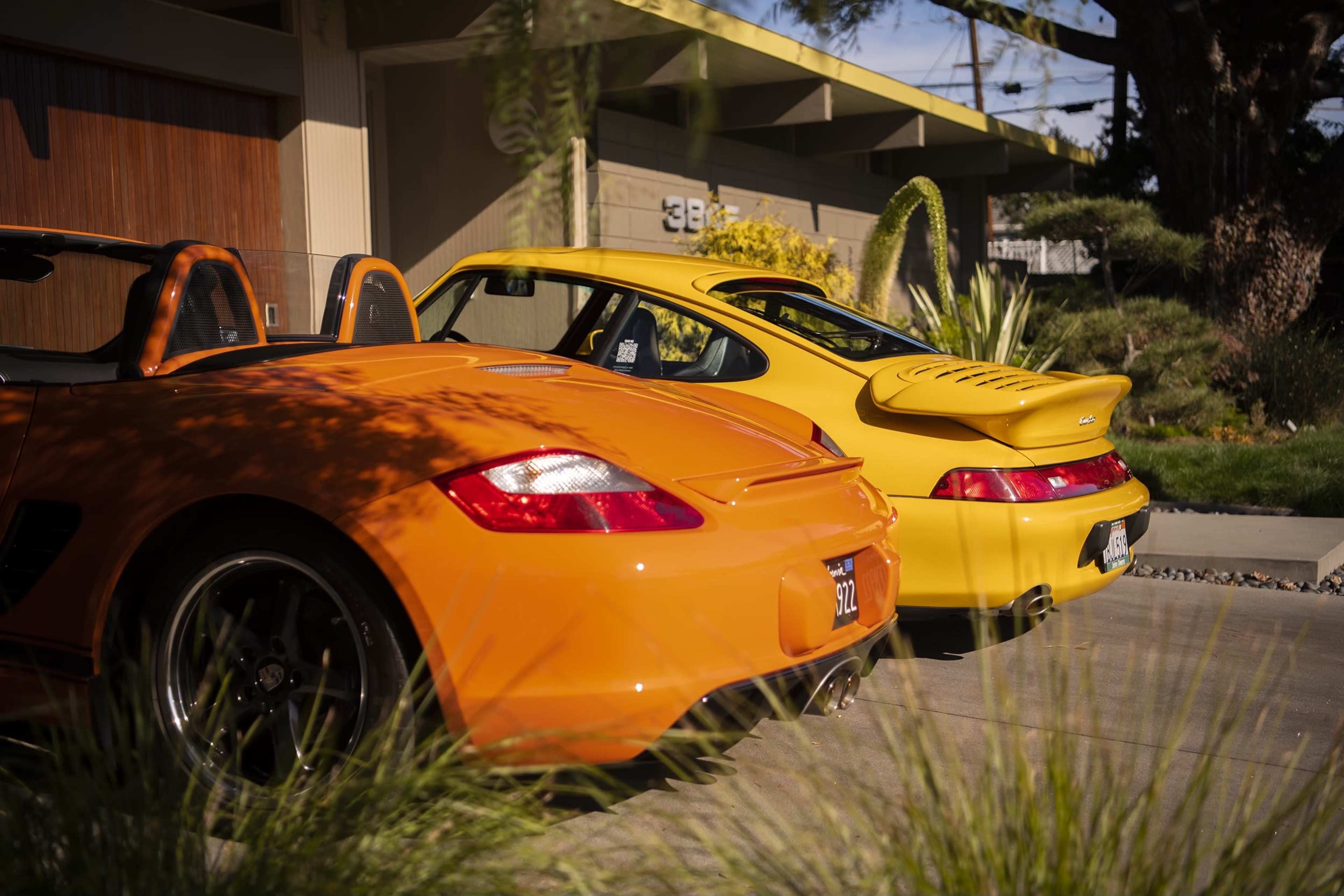 Two sports cars, one orange and one yellow, are parked side by side in a driveway surrounded by greenery.