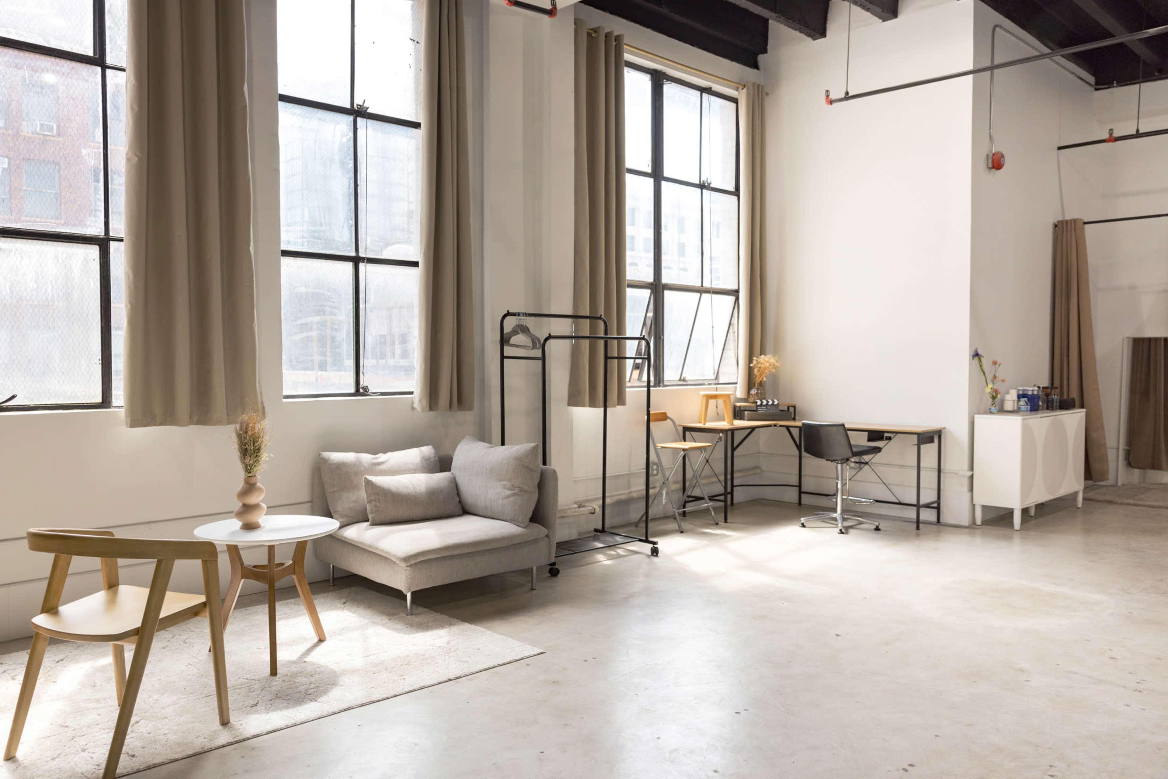 A spacious studio features large windows, minimalist furniture including a sofa and a table, and a workspace with a desk and chair.