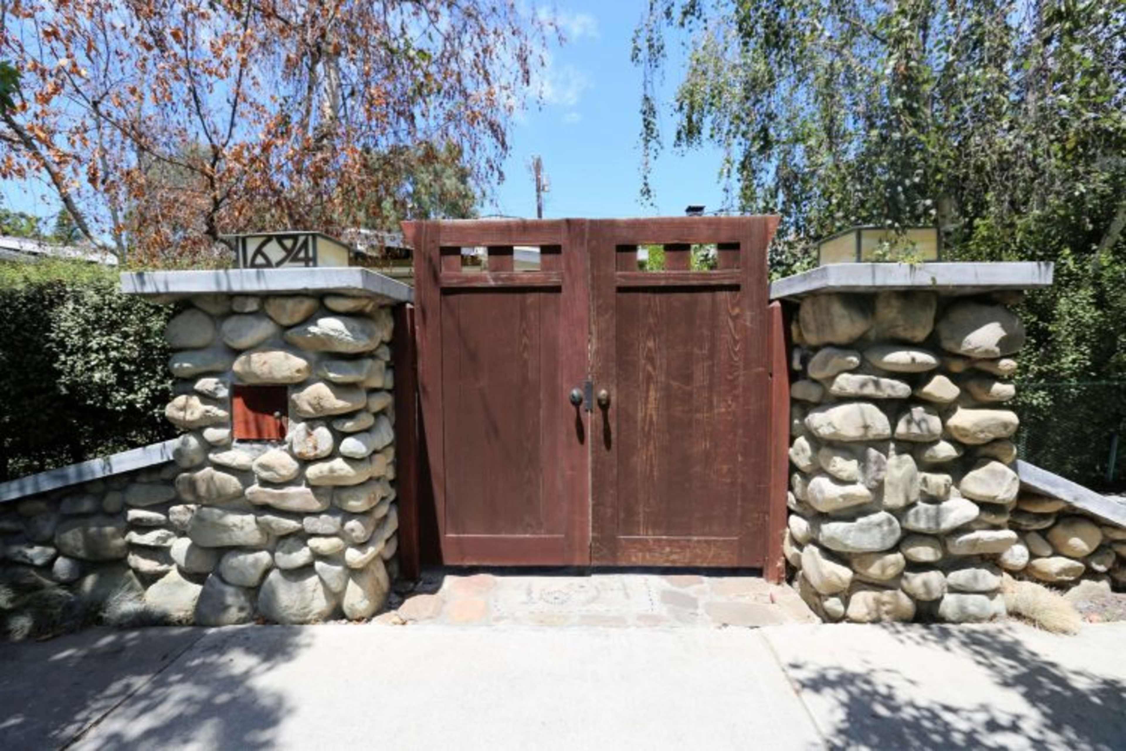 A wooden double gate with decorative panels stands between two stone pillars in a landscaped area.