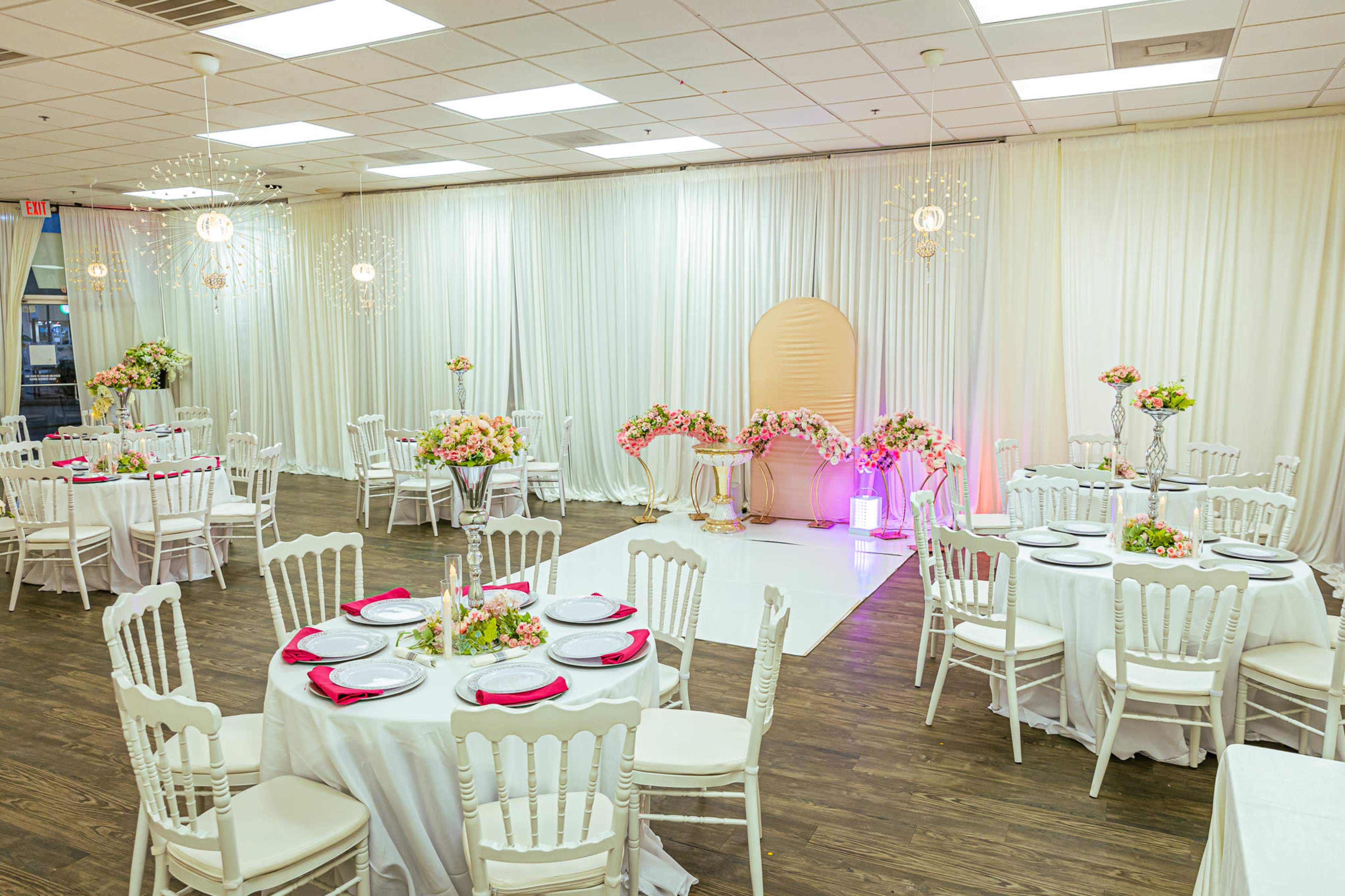 A banquet hall set up with round tables, each adorned with white tablecloths and pink accents, surrounded by floral arrangements and draped curtains.