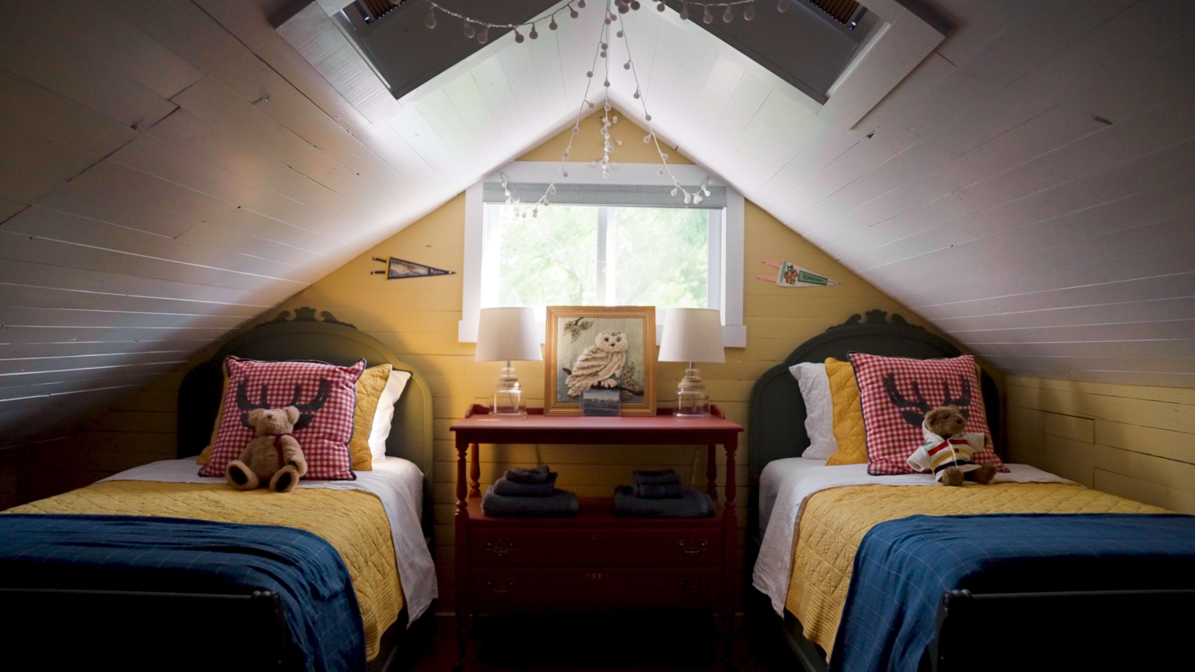 The image features a cozy attic bedroom with two twin beds, each adorned with checkered bedding, a shared nightstand, and decorative stuffed animals.
