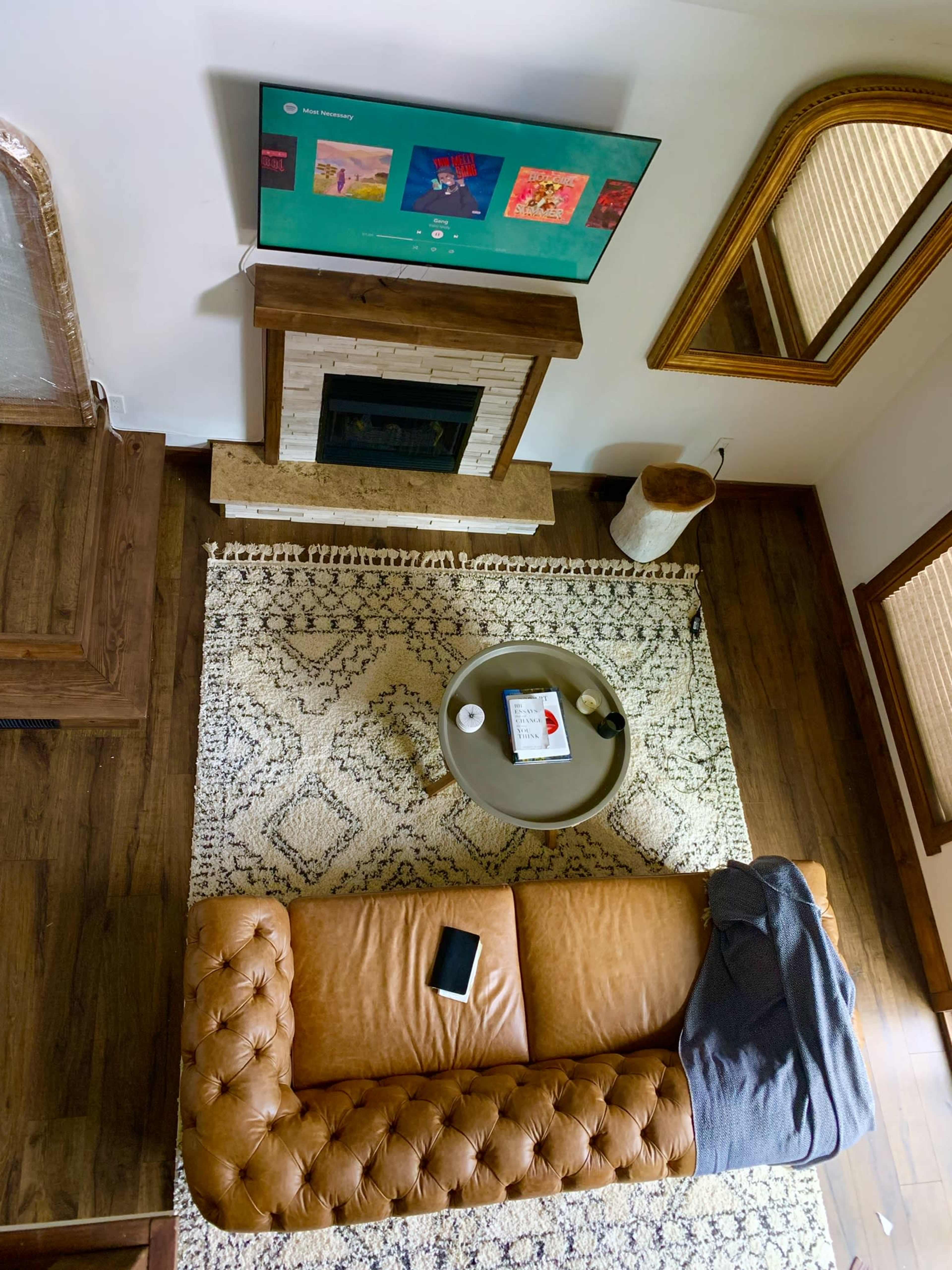 An overhead view of a living room featuring a leather couch, a round coffee table, a television mounted above a fireplace, and a decorative rug.