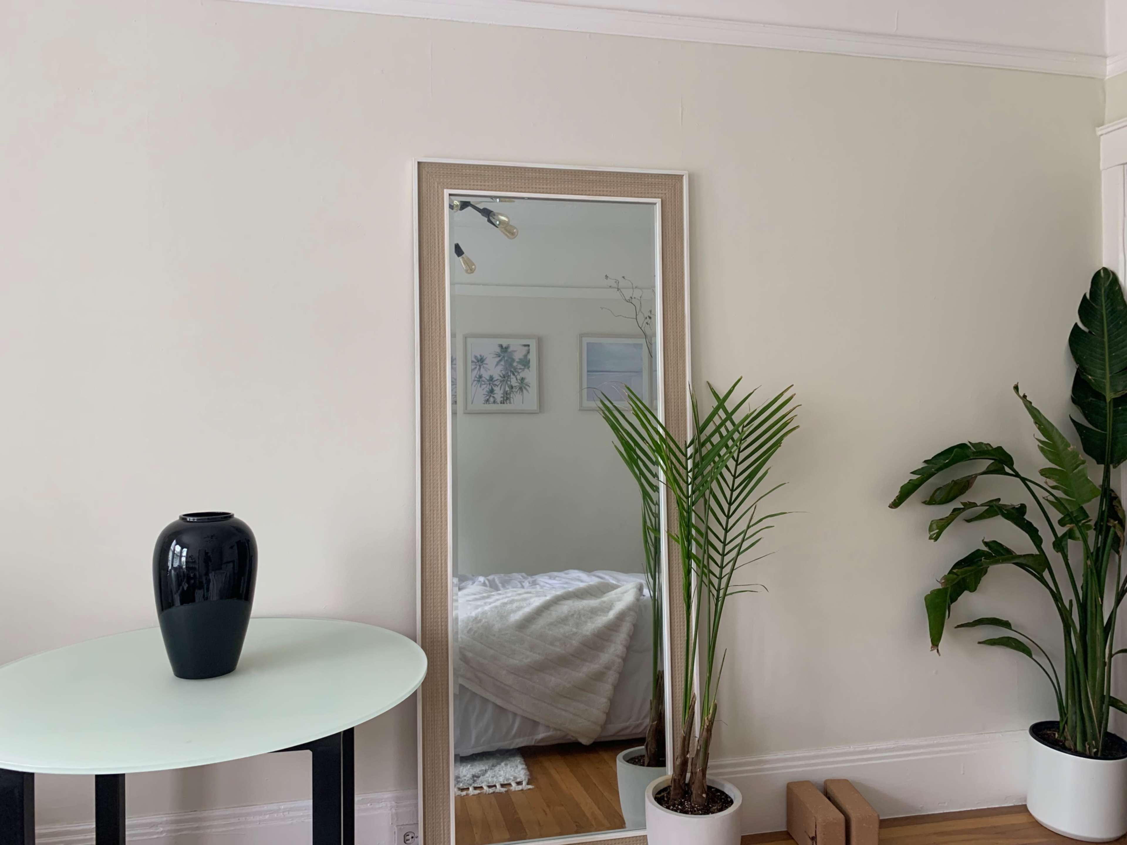 A decorative mirror hangs on a light-colored wall opposite a small table with a dark vase, alongside potted plants.