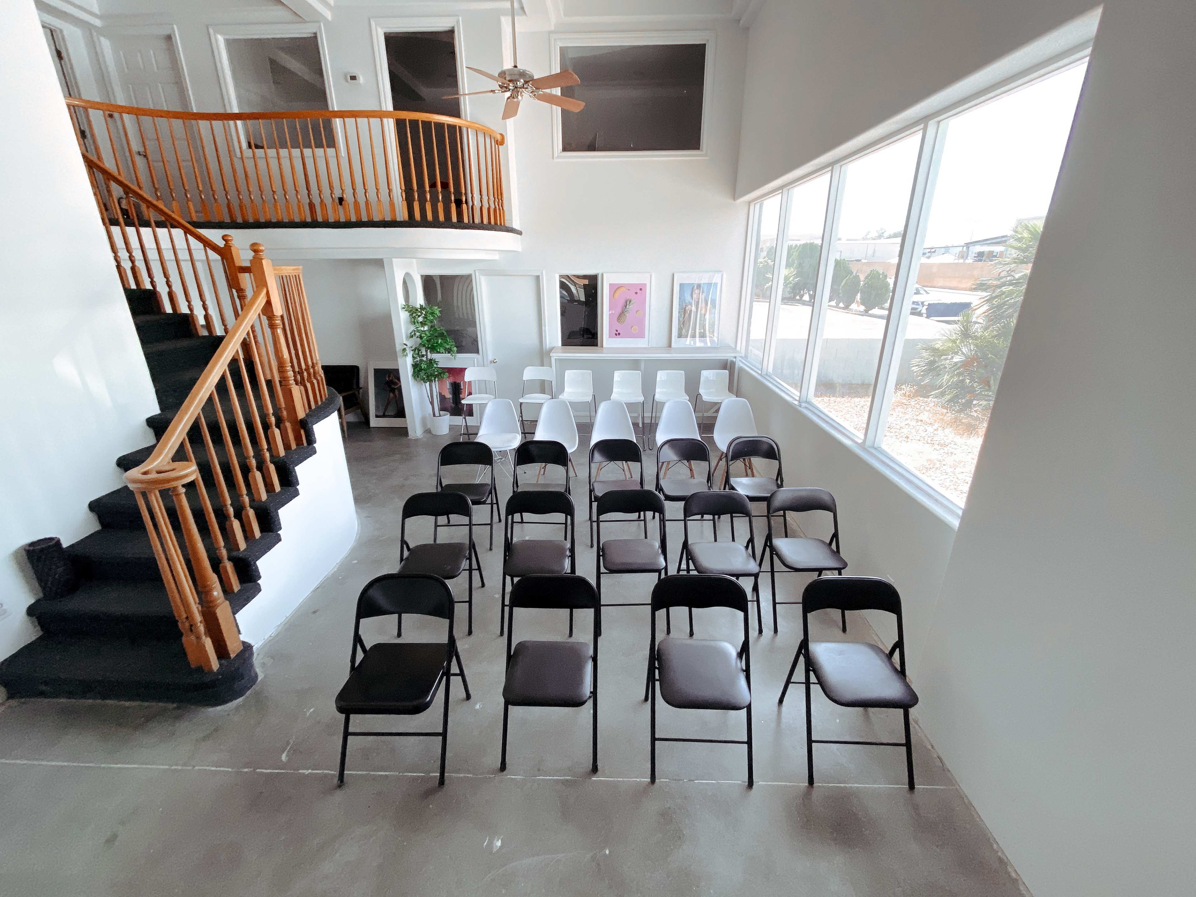 A row of black and white folding chairs is arranged in a spacious room with large windows and a staircase in the background.