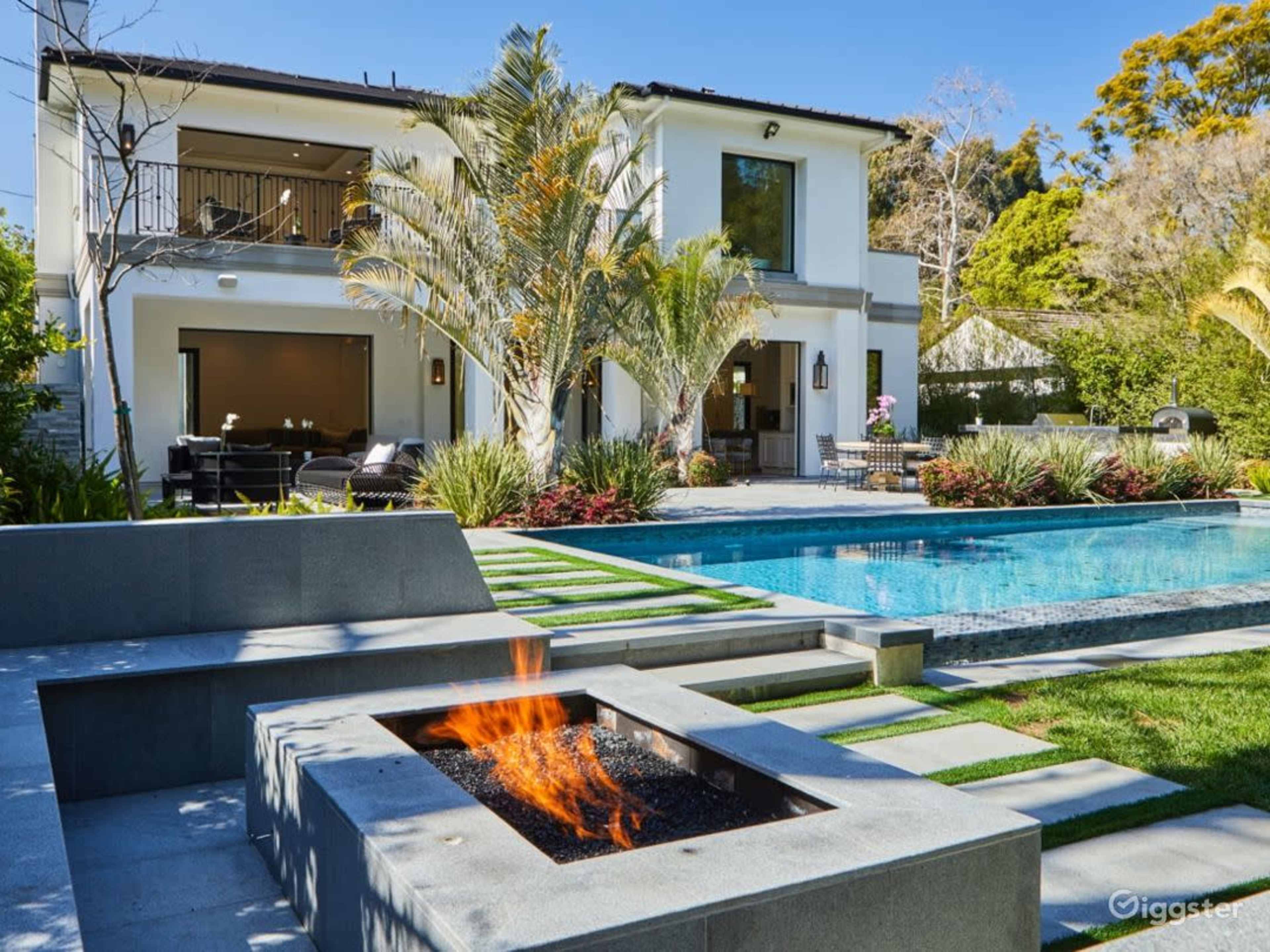 The image shows a modern two-story house with a spacious backyard featuring a pool, a fire pit, and landscaped greenery.