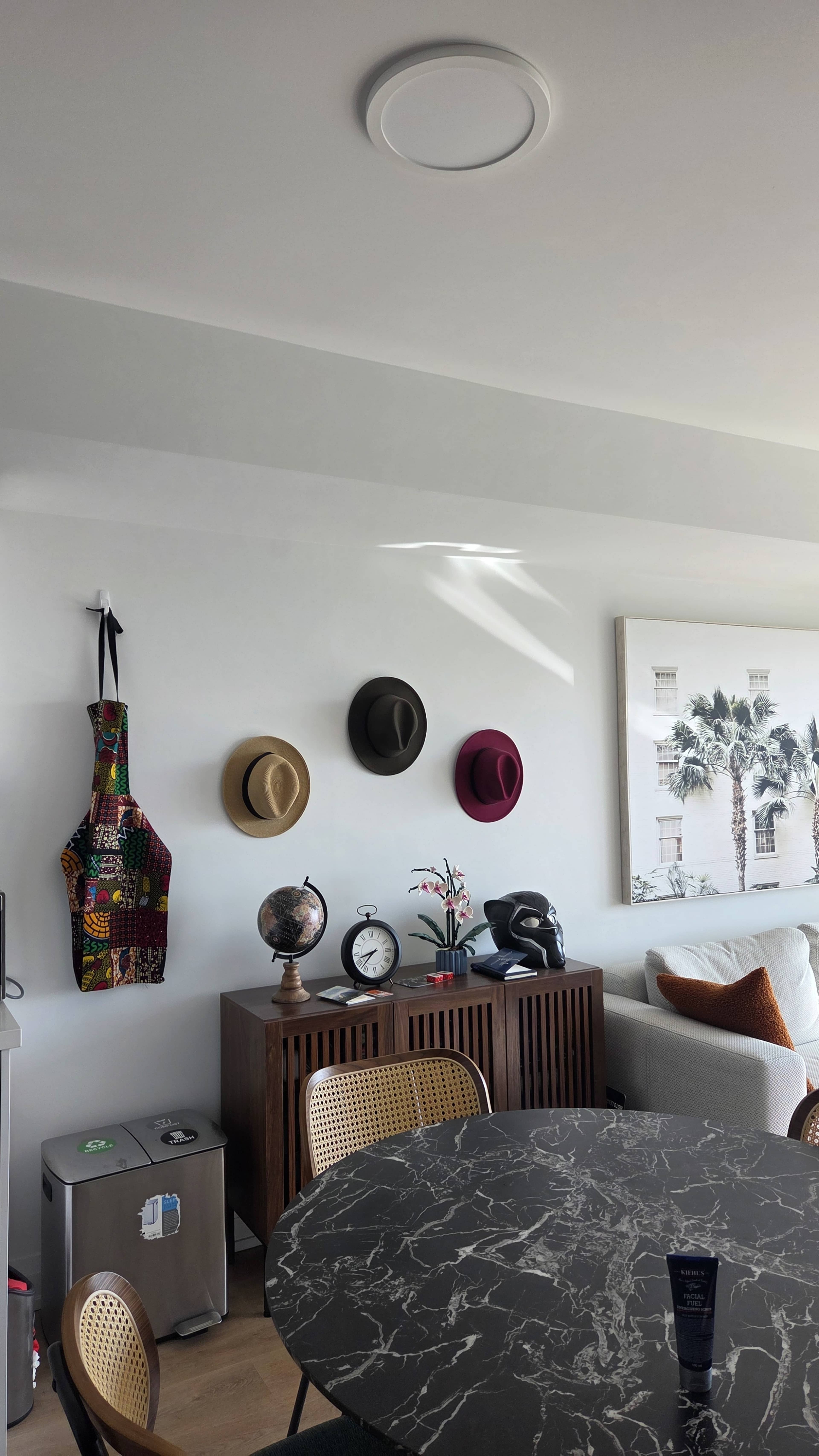 The image shows a modern living space with a round black marble dining table, a brown cabinet displaying a globe and clock, and three hats hanging on the wall above.