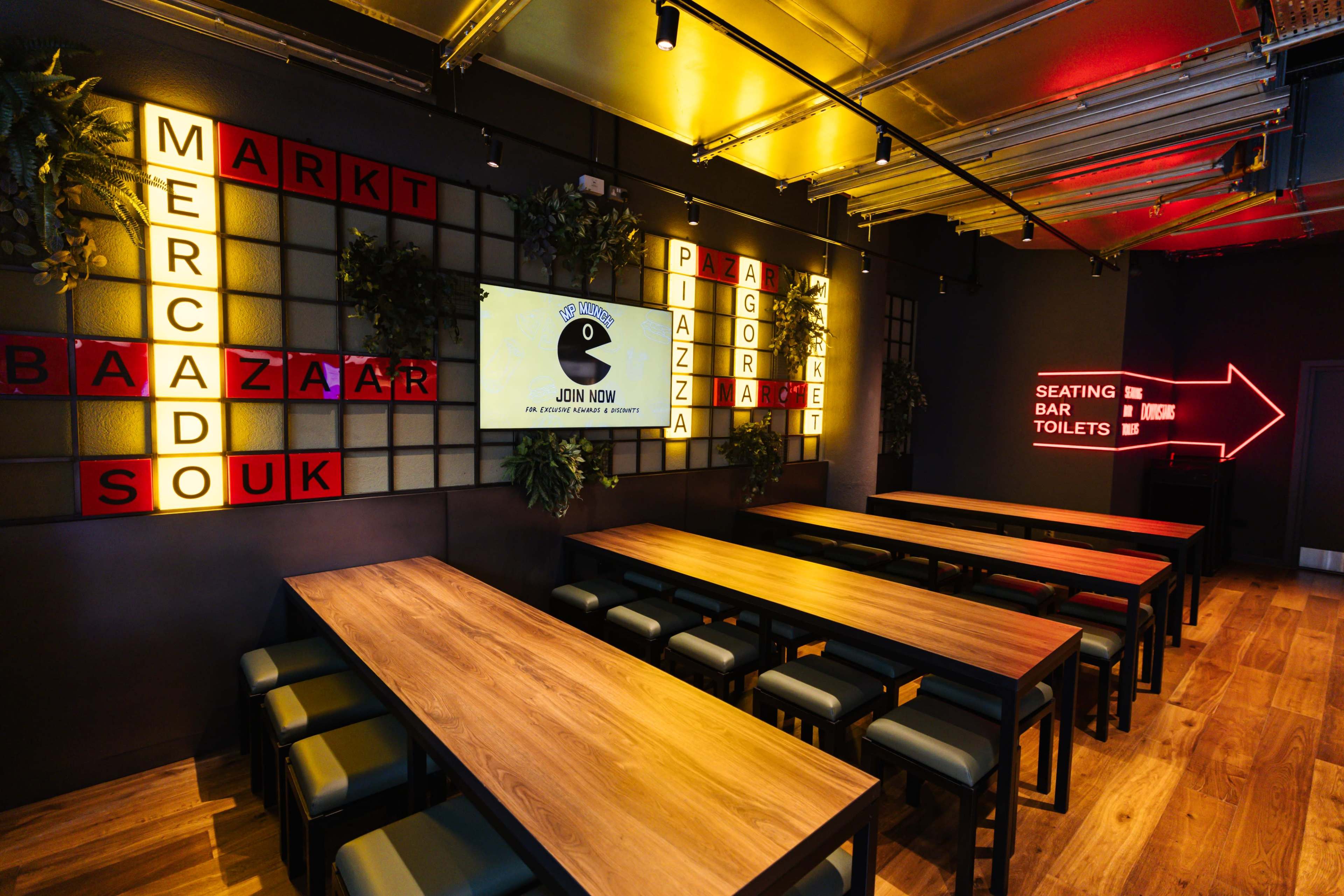 The interior of a dining space features wooden tables arranged in rows, with red and white signage on the walls indicating various areas such as "SEATING," "BAR," and "TOILETS."
