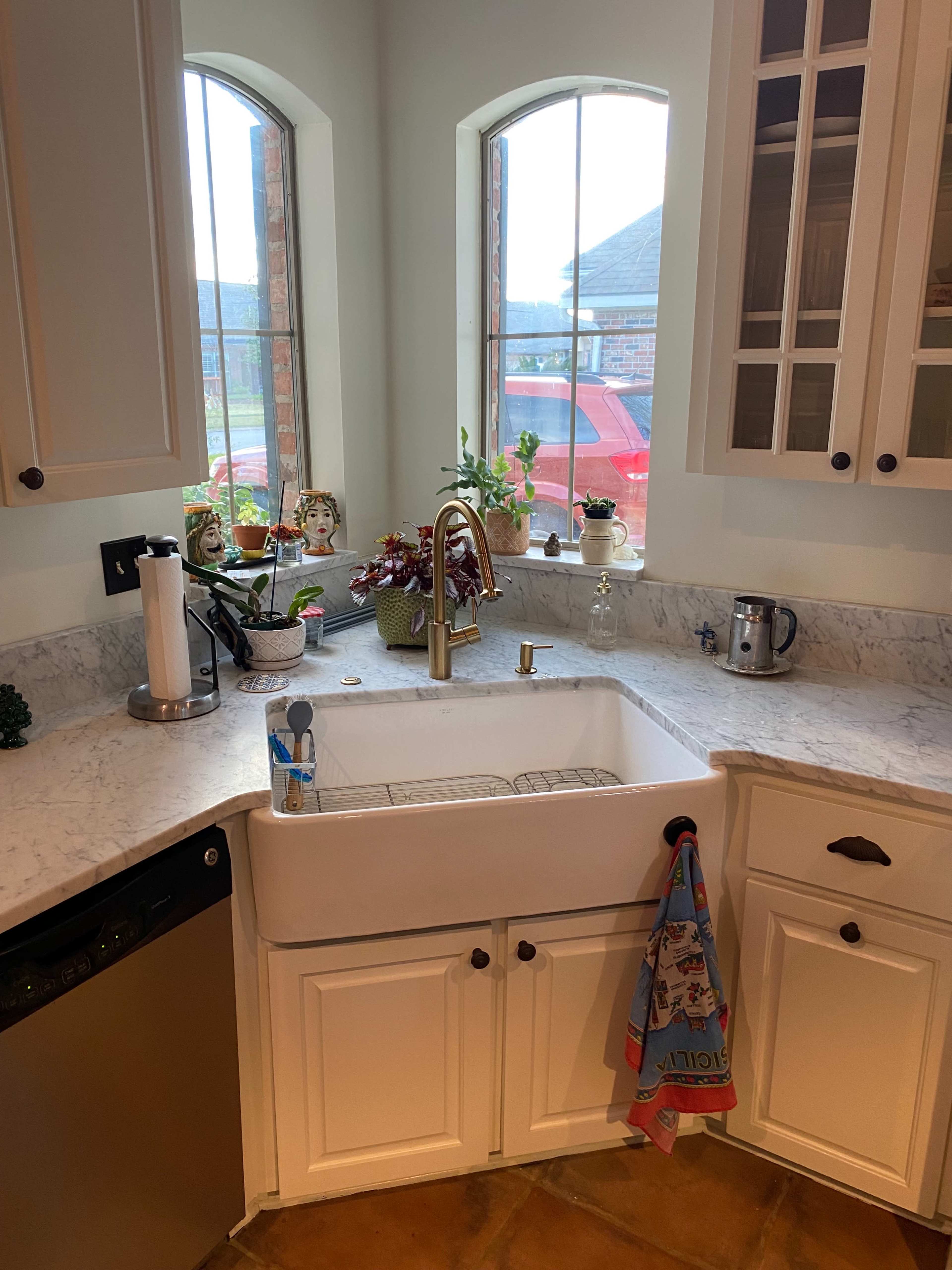 A kitchen features a farmhouse sink set into a marble countertop with two windows above, displaying plants and a red car outside.