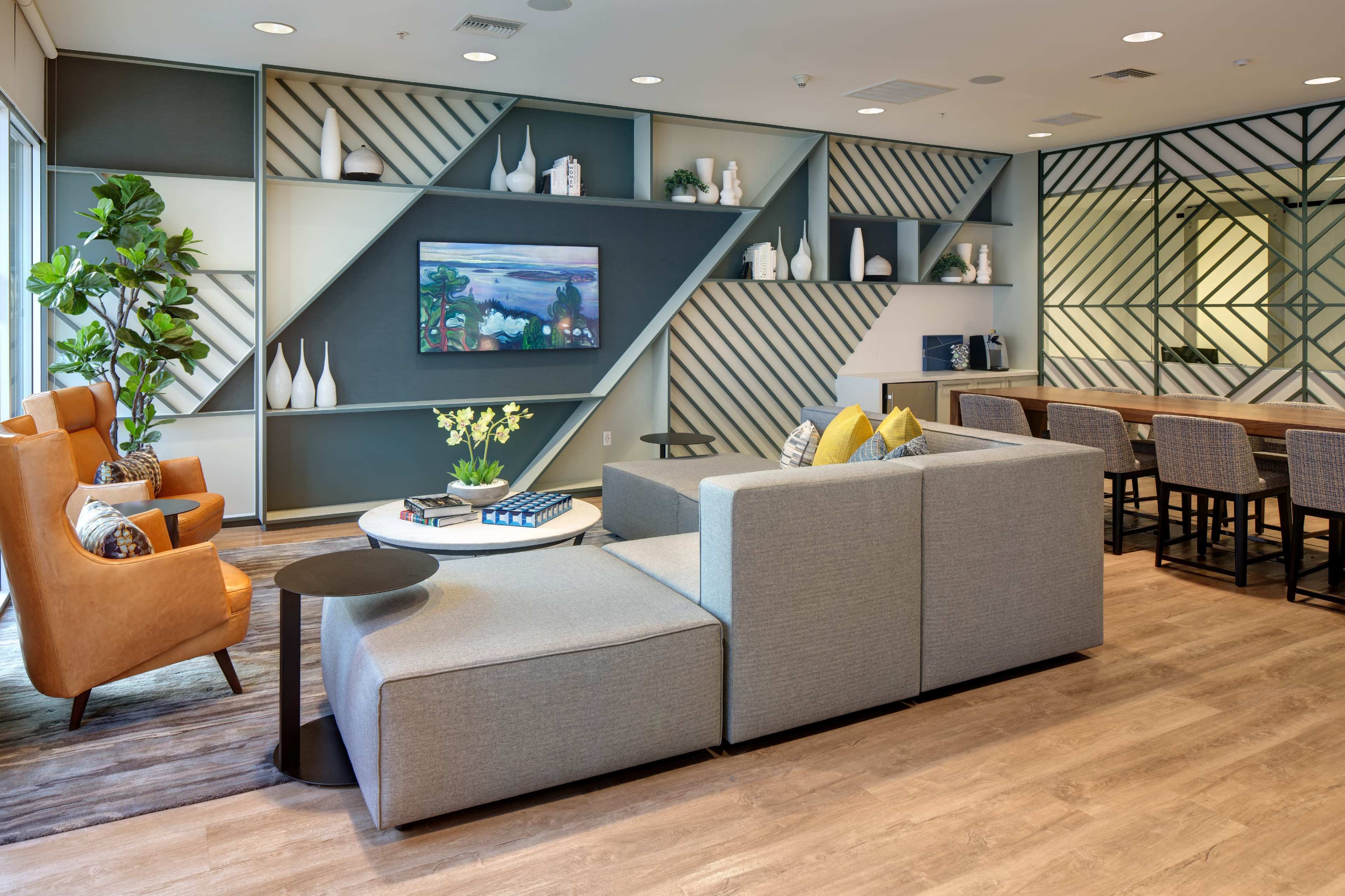 A modern lounge area featuring a sectional sofa, orange armchairs, a round coffee table with decorative items, and a long table with high chairs against a patterned wall.