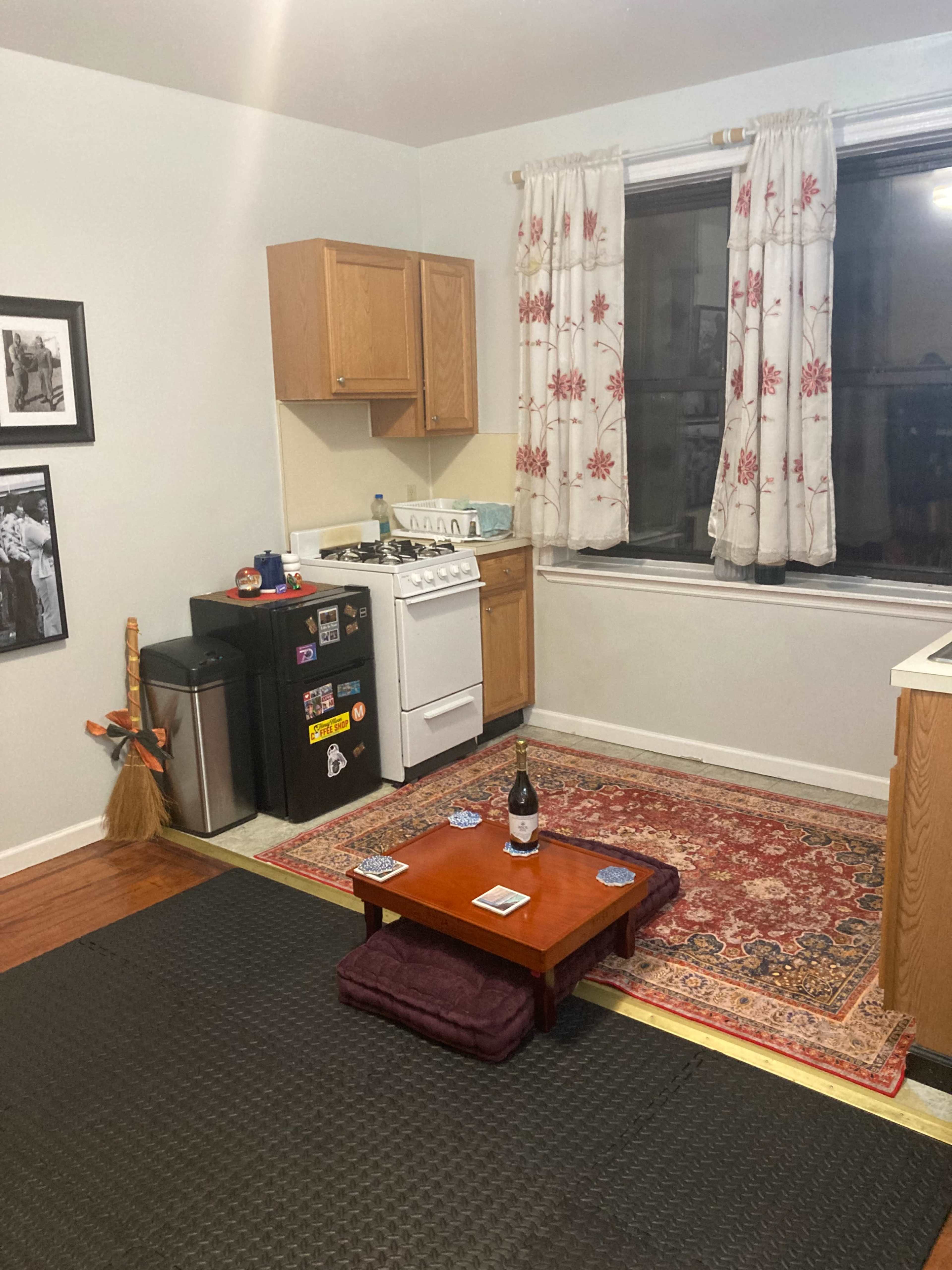 The image shows a small kitchen area with wooden cabinets, a white stove, a black refrigerator, and a red rug on the floor beneath a low table surrounded by cushions.