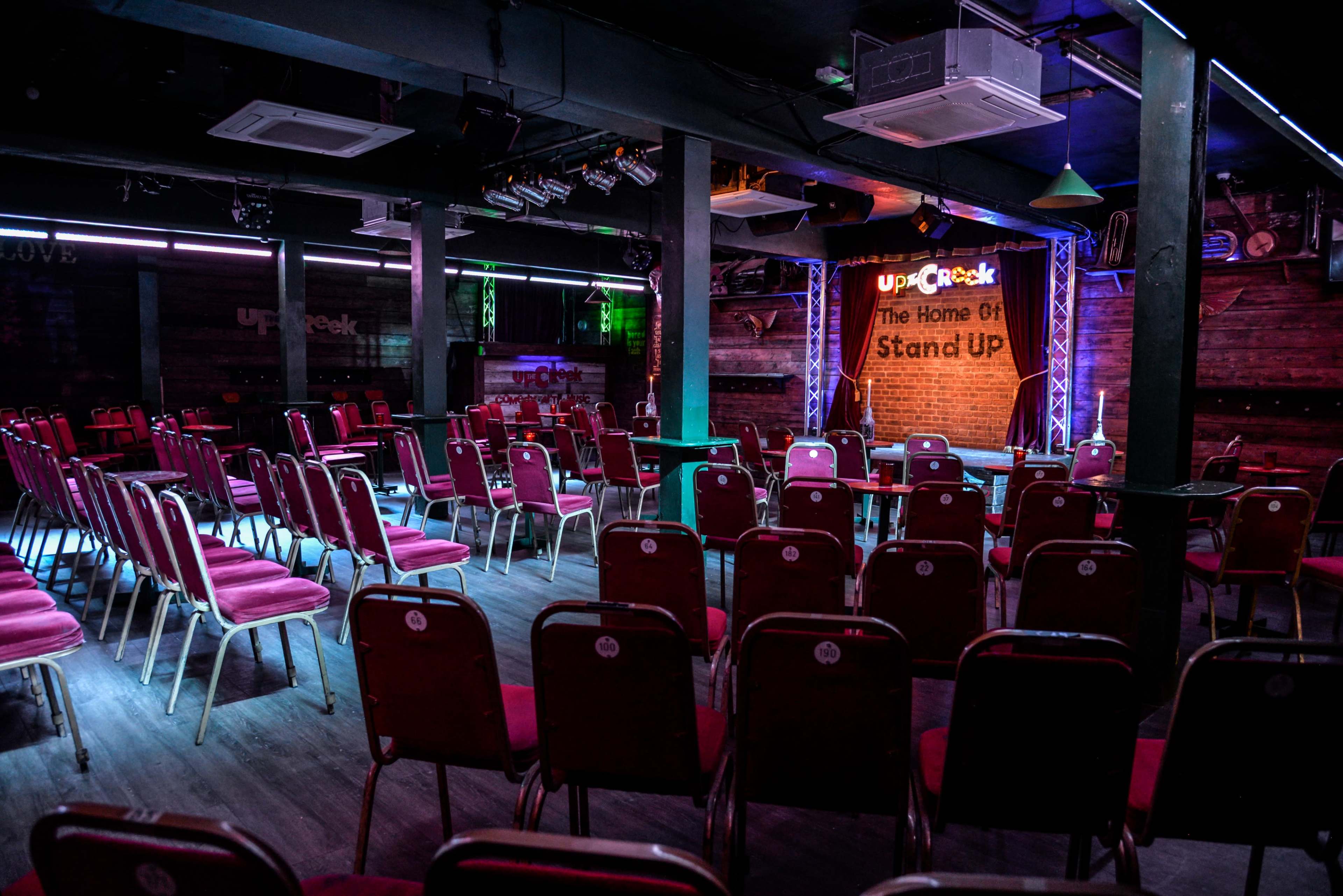 The image shows a comedy club stage with a brick backdrop, empty chairs arranged in rows facing the stage, and dim lighting.