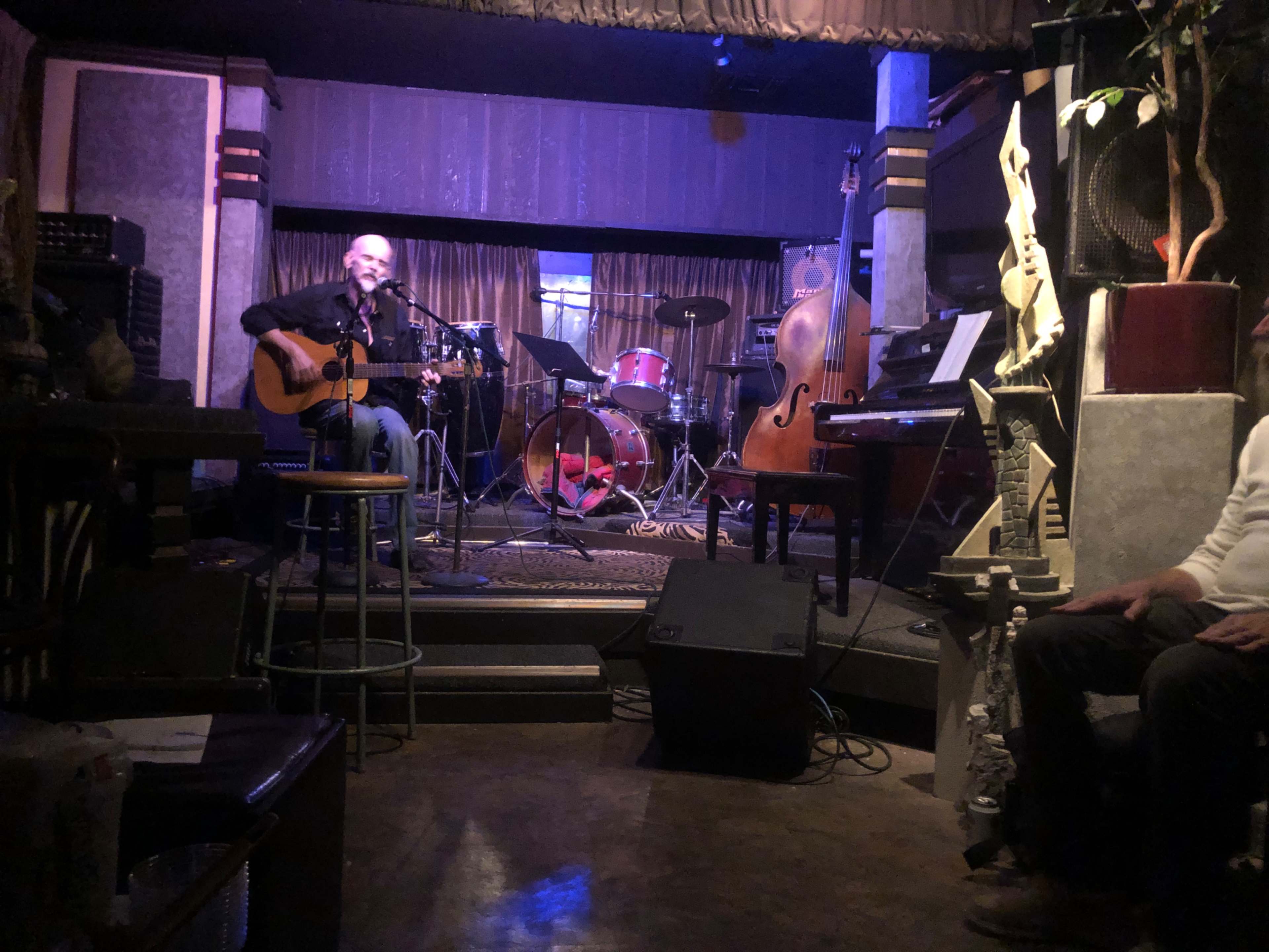 A musician plays guitar and sings on stage in a cozy music venue, surrounded by various instruments and a small audience.