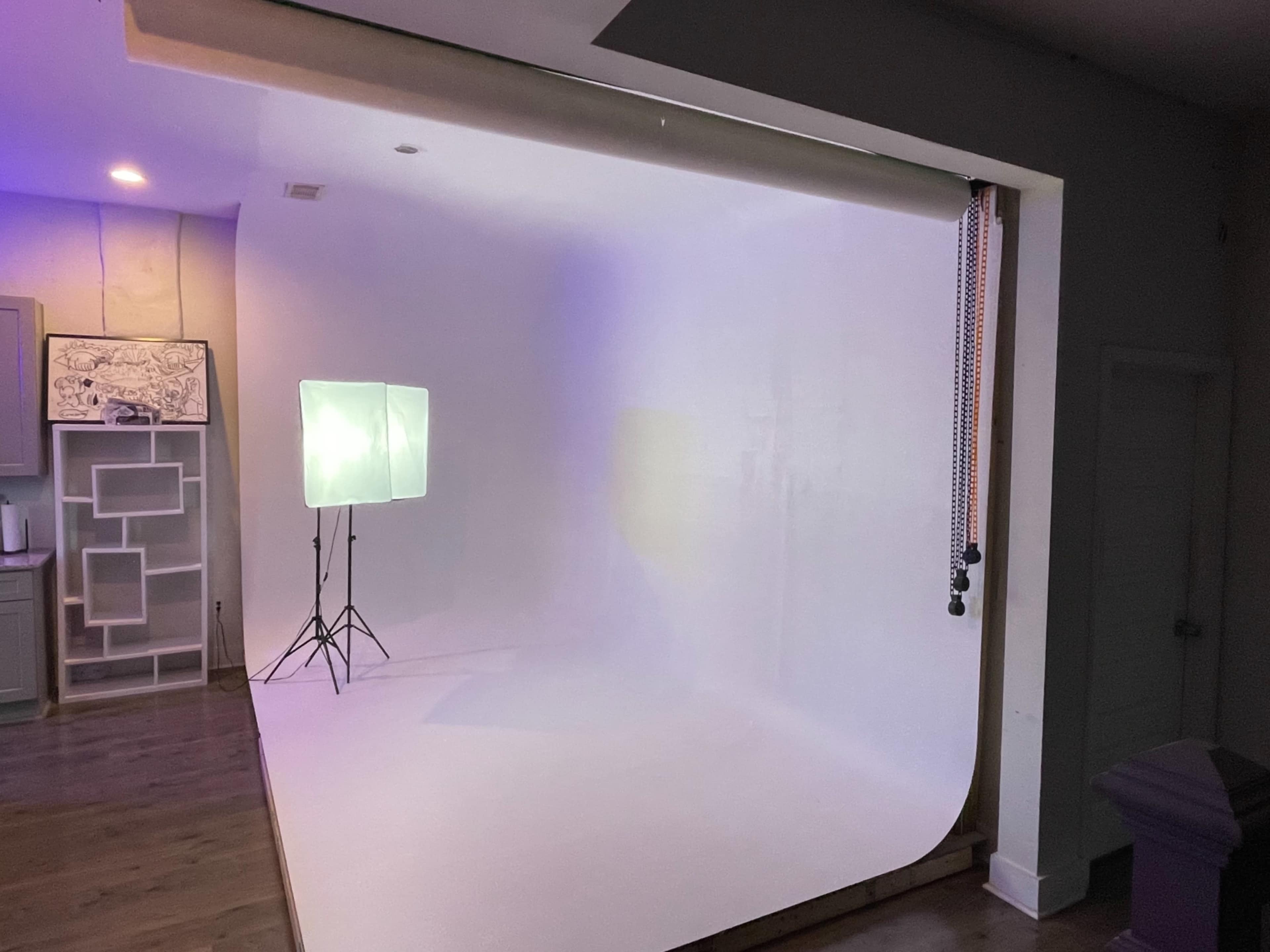 A well-lit photography studio featuring a seamless white backdrop and a softbox light set up on a tripod.