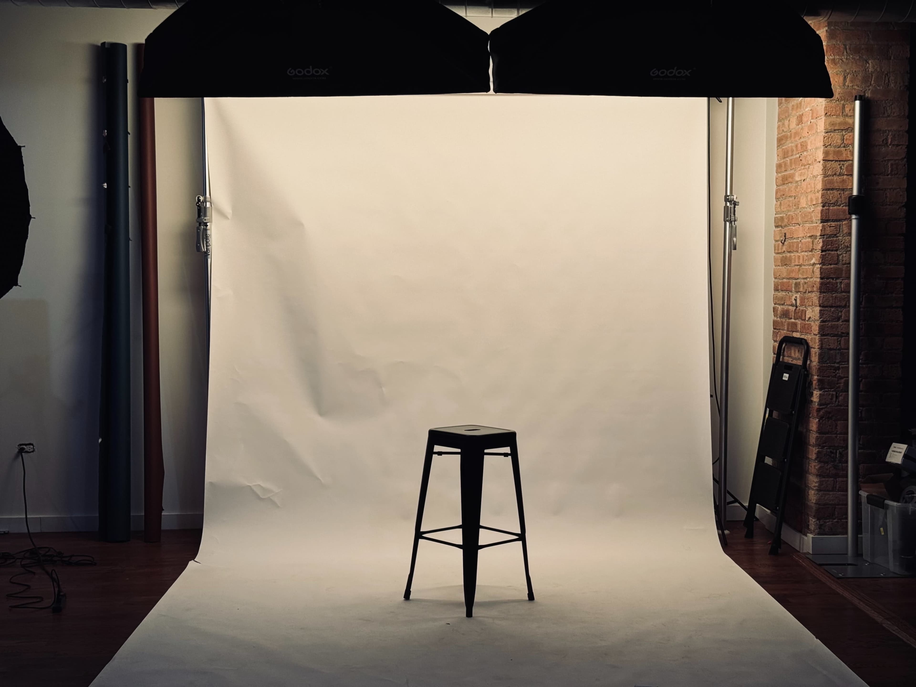 A black metal stool is positioned in front of a white backdrop illuminated by two studio lights.