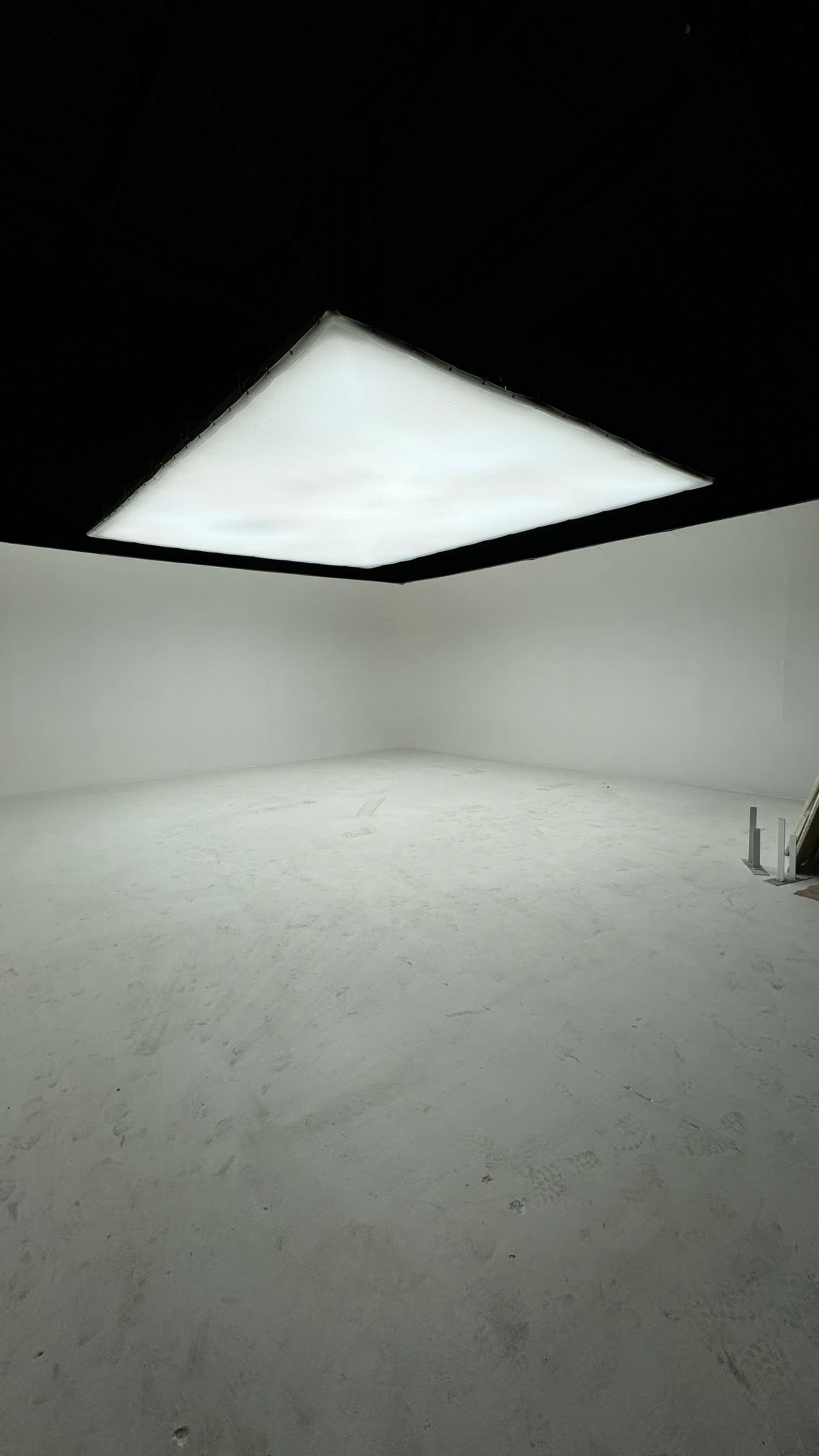 A large, flat overhead light illuminates an empty room with white walls and a bare floor.